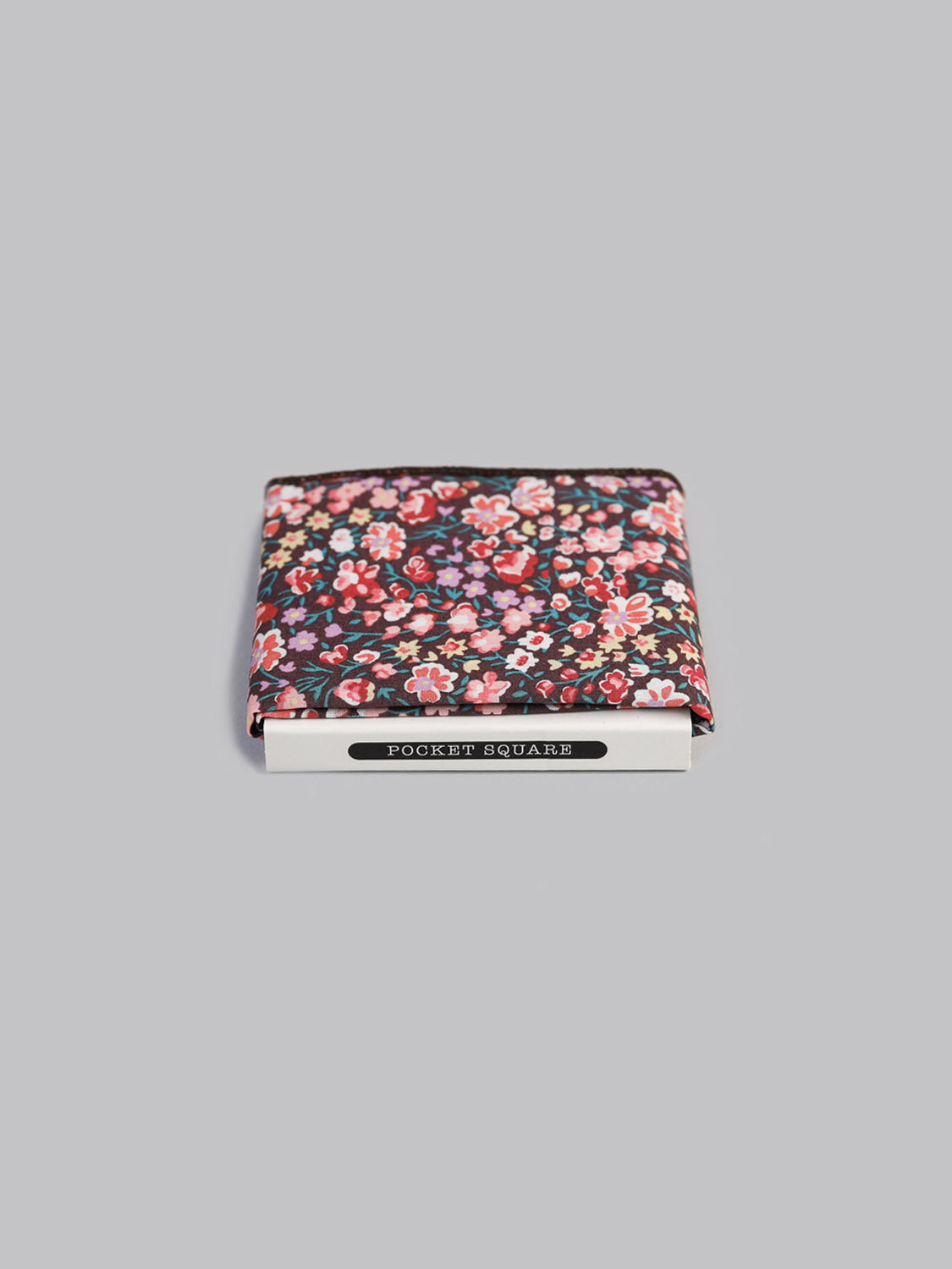 A folded Liberty™ Jo Pocket Square by ID Menswear, featuring a red and pink floral print on black Tana Lawn cotton, is partially packaged in a white “Pocket Square” box against a plain grey background.
