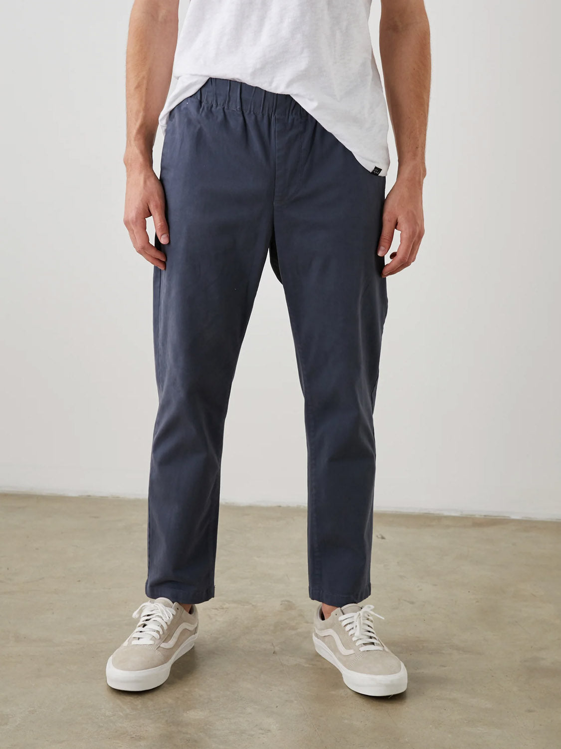 A person wearing a white t-shirt and Rails Julian Pant by Rails in navy blue with an elastic waistband stands on a concrete floor, pairing the vintage-look pants with stylish white sneakers indoors.