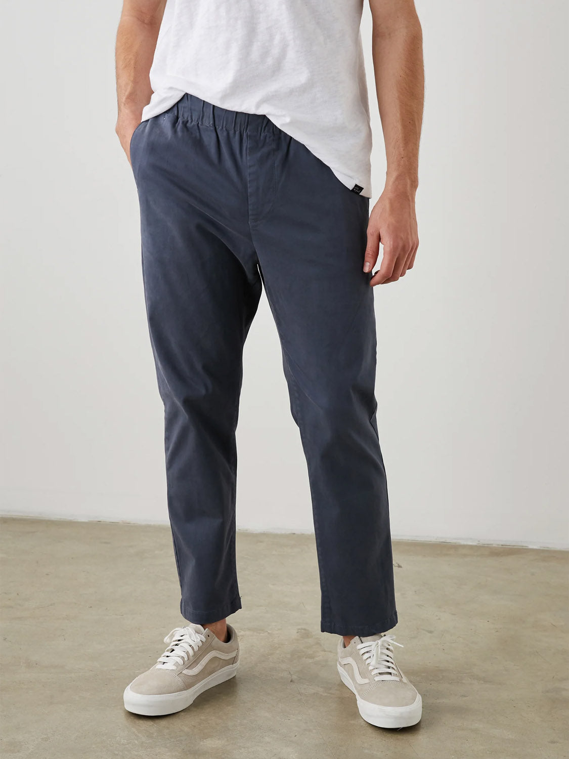 A person wearing a white t-shirt and Rails Julian Pant in navy blue stands with hands in pockets, complemented by light-colored sneakers. The background features a light gray wall and a neutral-toned floor.