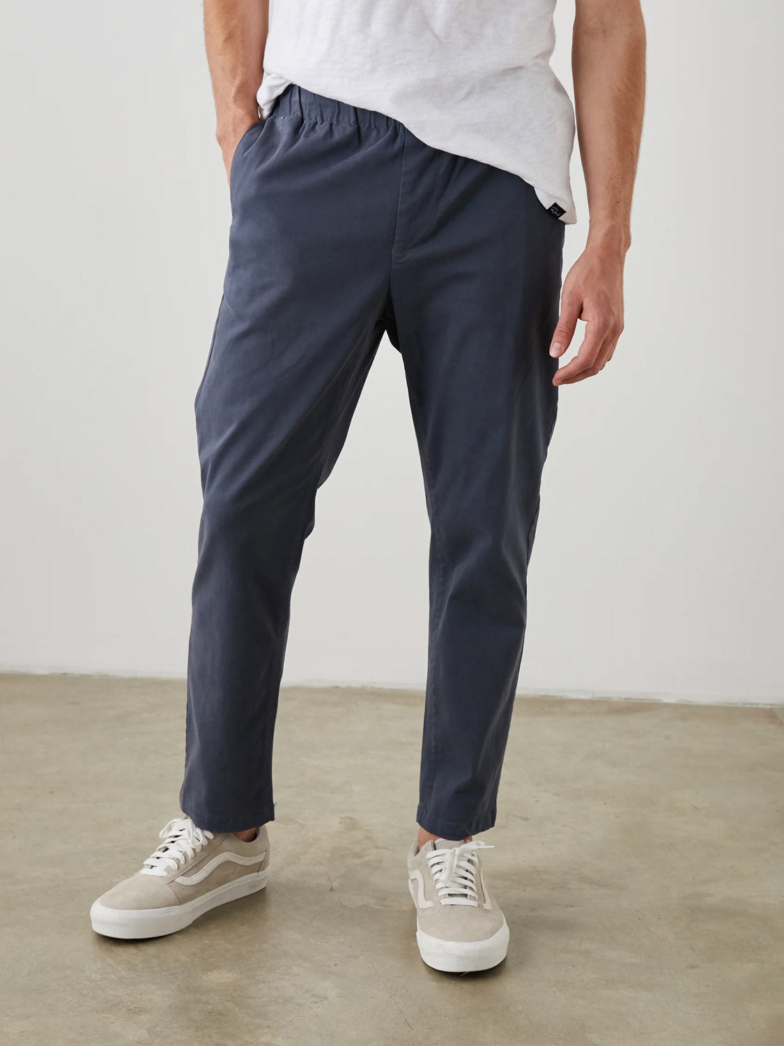 A person wearing a white T-shirt and Rails Julian Pant stands against a plain white wall. Hands in pockets, they wear beige and white sneakers. The light brown concrete floor adds an earthy touch to their casual style.