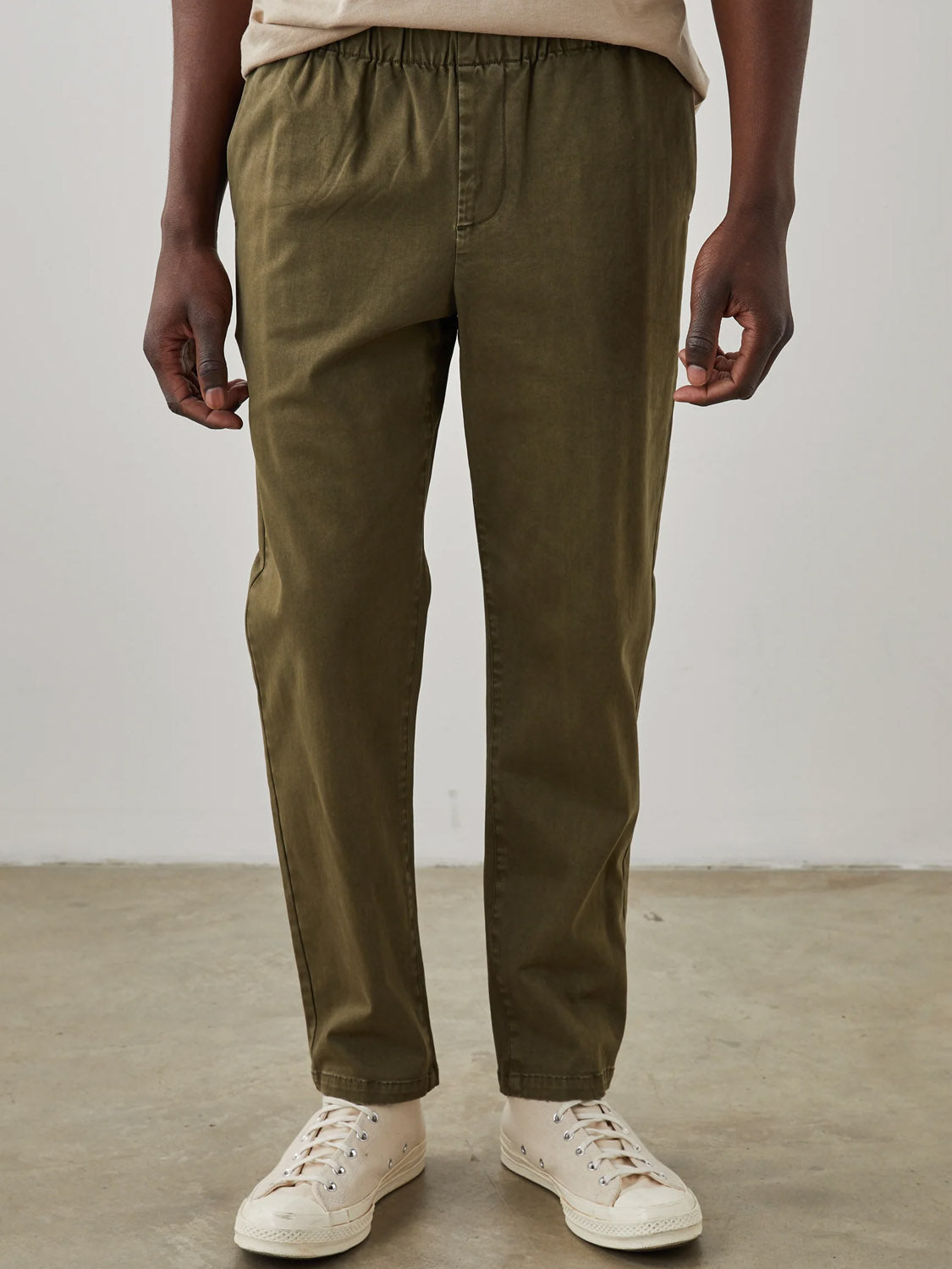 Wearing Rails Julian Pant in olive green stretch twill and white sneakers, a person stands on a concrete floor against a plain white background. Their relaxed hands by their sides give off a casual yet stylish vibe.