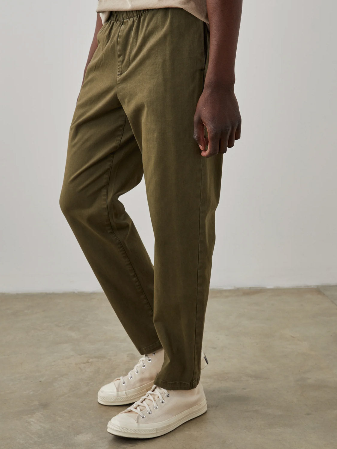 On a concrete floor, someone models the Rails Julian Pant from Rails. The olive green, elastic waist pants offer a relaxed fit and comfort with their stretch twill fabric, paired effortlessly with white sneakers. The style exudes a vintage vibe against a plain, light-colored wall backdrop.