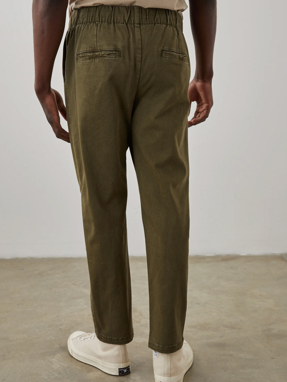 Standing on concrete, a person faces away in olive green Rails Julian Pants with a vintage vibe. The stretch twill design includes an elastic waistband and back pockets, complemented by white sneakers against a plain, light-colored wall.