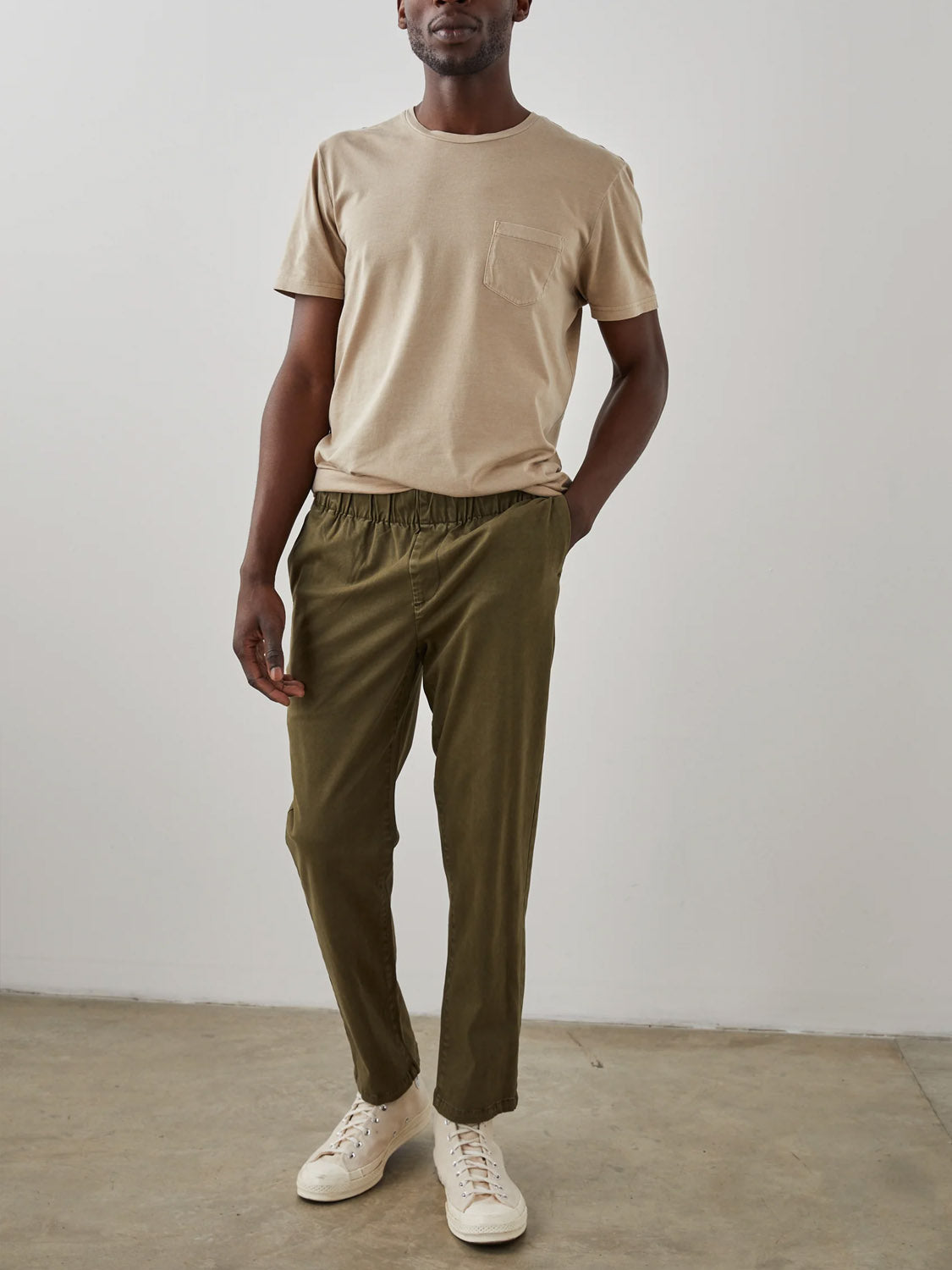 A man stands casually with one hand in his pocket against a white wall and concrete floor, wearing a beige t-shirt, Rails Julian Pant in vintage-look olive green by Rails, and white sneakers.