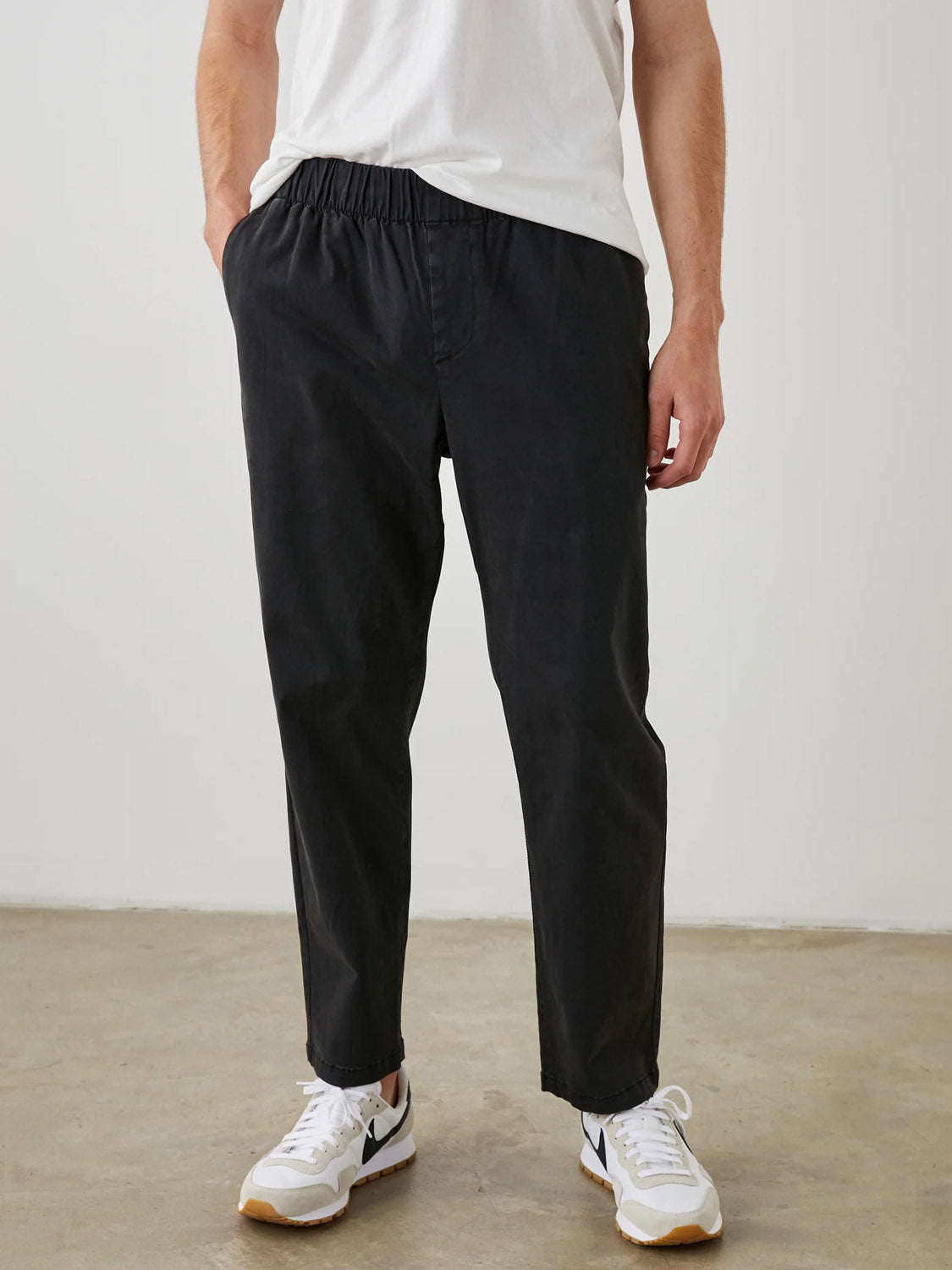 A person wearing black Rails Julian Pant, a white t-shirt, and white sneakers with black accents stands casually with one hand in their pocket against a plain background.