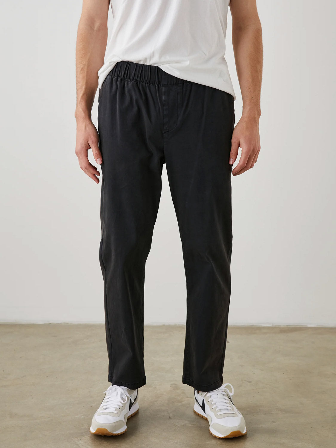 A person wearing a plain white t-shirt, Rails Julian Pant with an elastic waistband, and white sneakers stands on a concrete floor against a plain white background. The focus is on the lower half of the body.