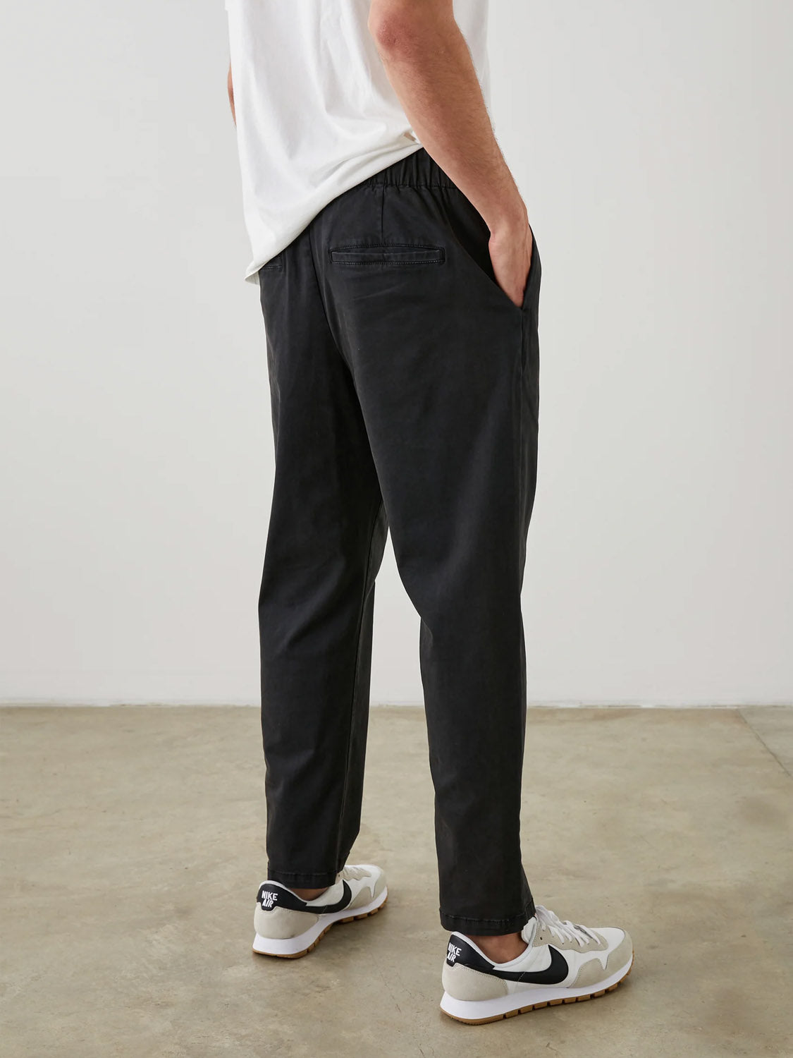 A person in a white shirt and Rails Julian Pant stands with hands in pockets, facing away. They wear black and white sneakers. The gray concrete floor complements the plain white wall behind them.