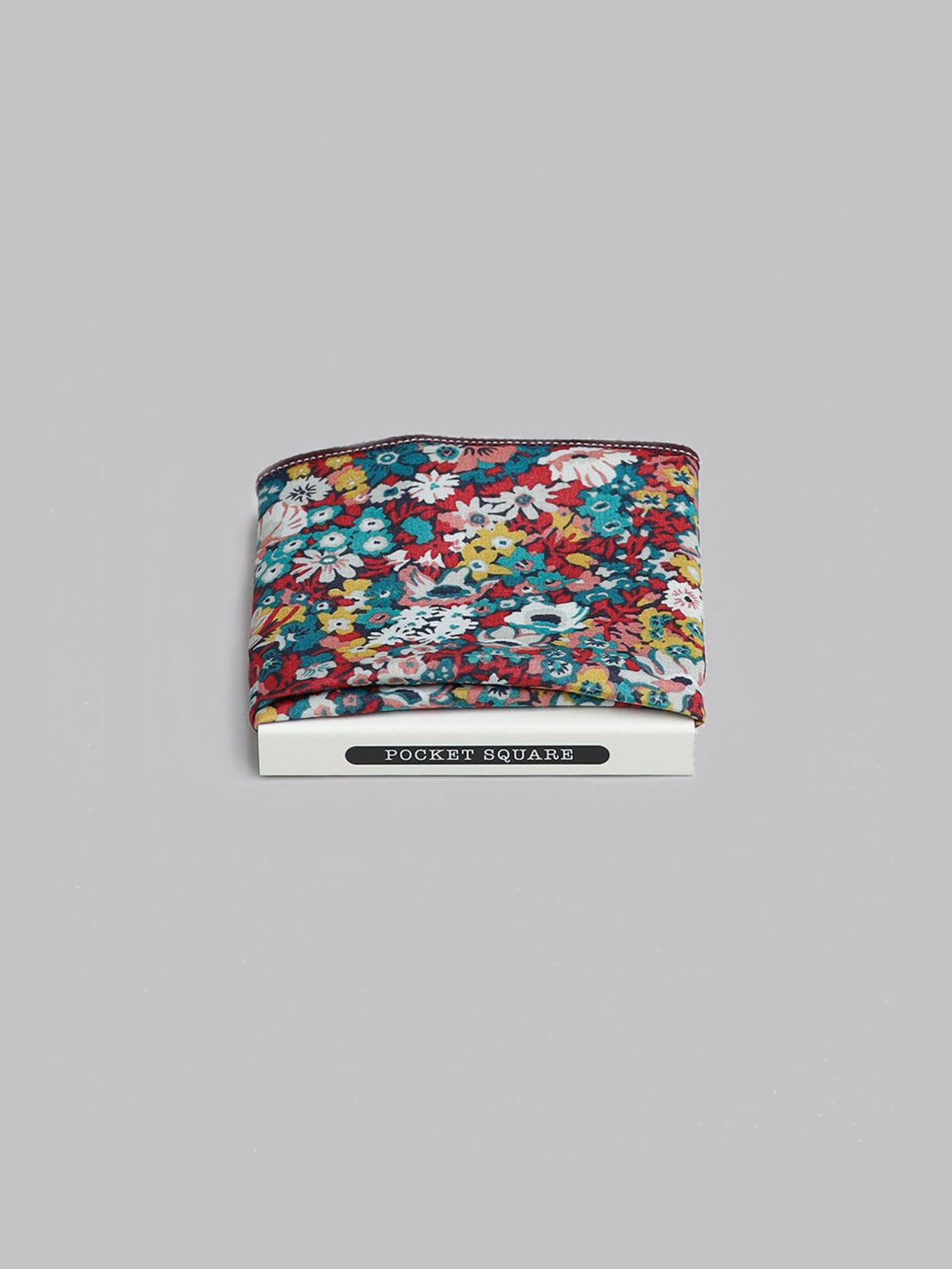 A folded ID Menswear Liberty™ June Pocket Square, handmade in Brooklyn from soft Tana Lawn cotton, displays a vibrant red, blue, yellow, and white floral pattern on light grey. The POCKET SQUARE label is visible at the bottom.