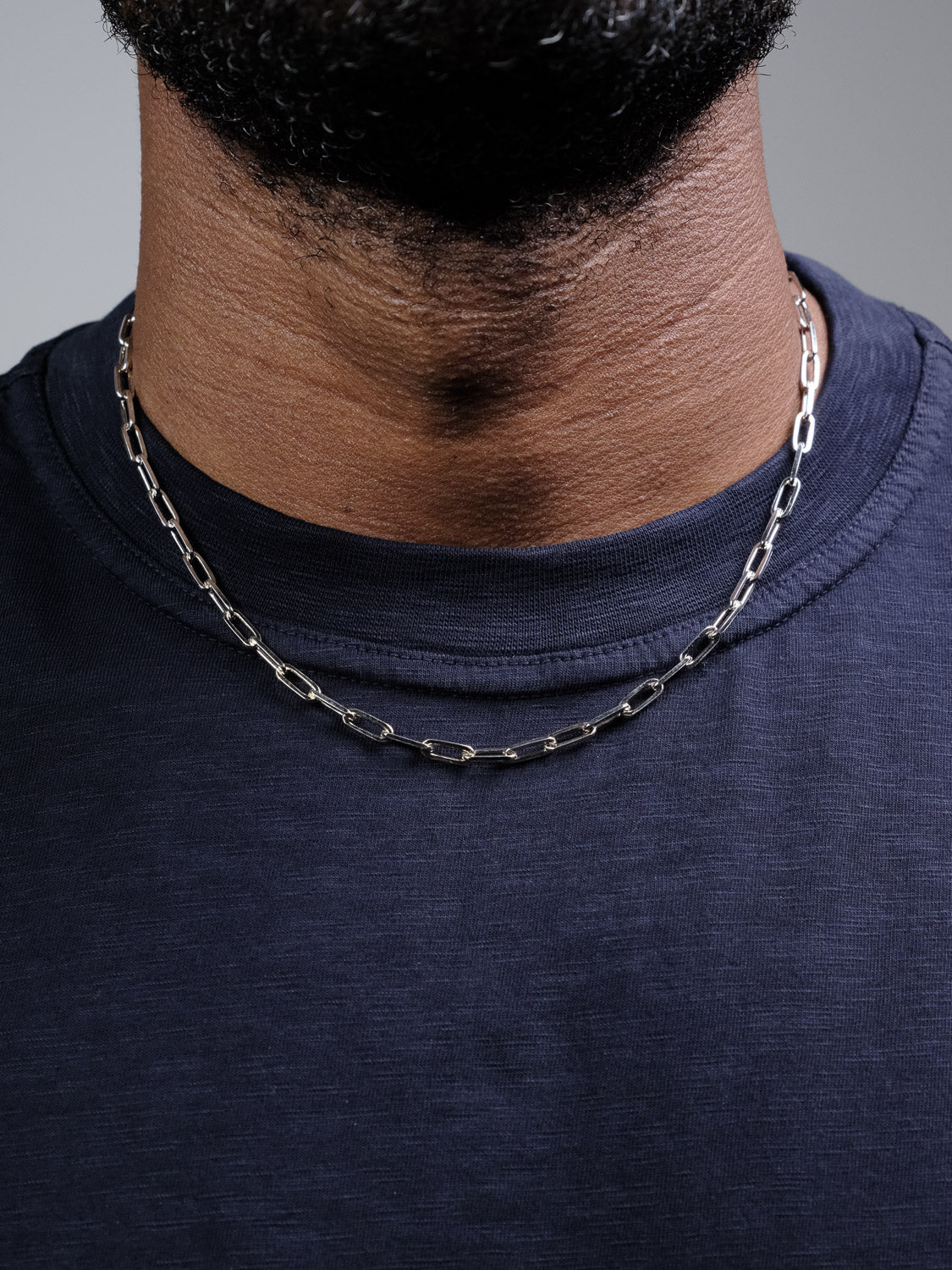 A close-up of a person's neck wearing the Kai 120 Paper Clip Chain Necklace by ID Menswear over a navy blue t-shirt, focusing on the necklace and shirt neckline, with the lower part of the face not shown.