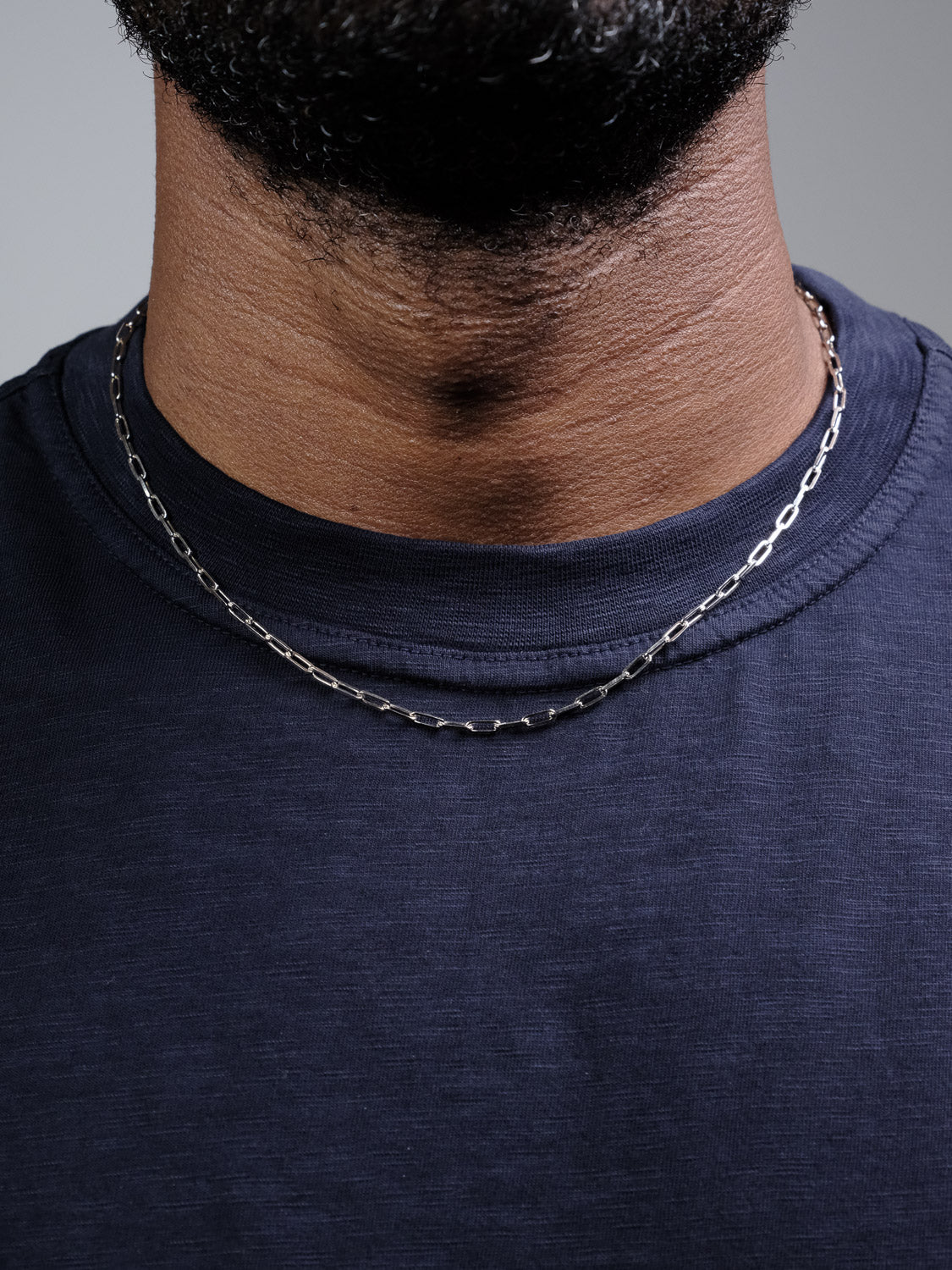 Close-up of a person wearing the Kai 80 Paper Clip Necklace by ID Menswear with a dark blue crew-neck shirt. Only the lower part of their face, neck, and upper chest are shown.
