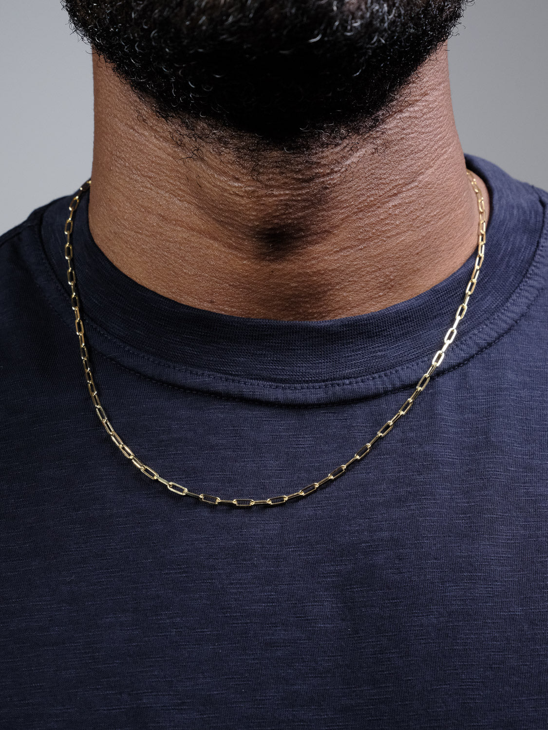 A close-up shows a person wearing the Kai 80 Paper Clip Necklace by ID Menswear, featuring an elongated gold-plated link design, paired with a navy blue crew-neck shirt against a neutral background.