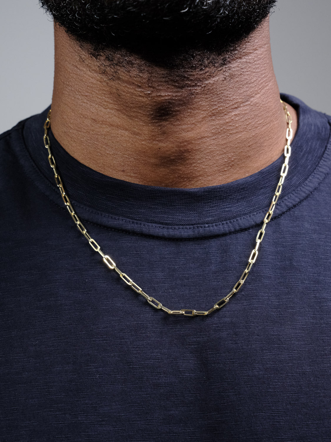 A close-up of a person wearing a navy blue shirt and the Kai 100 Paper Clip Chain Necklace by ID Menswear, a gold plated necklace with elongated links made in Italy. The focus is on their neck and upper chest, highlighting the elegant chain.