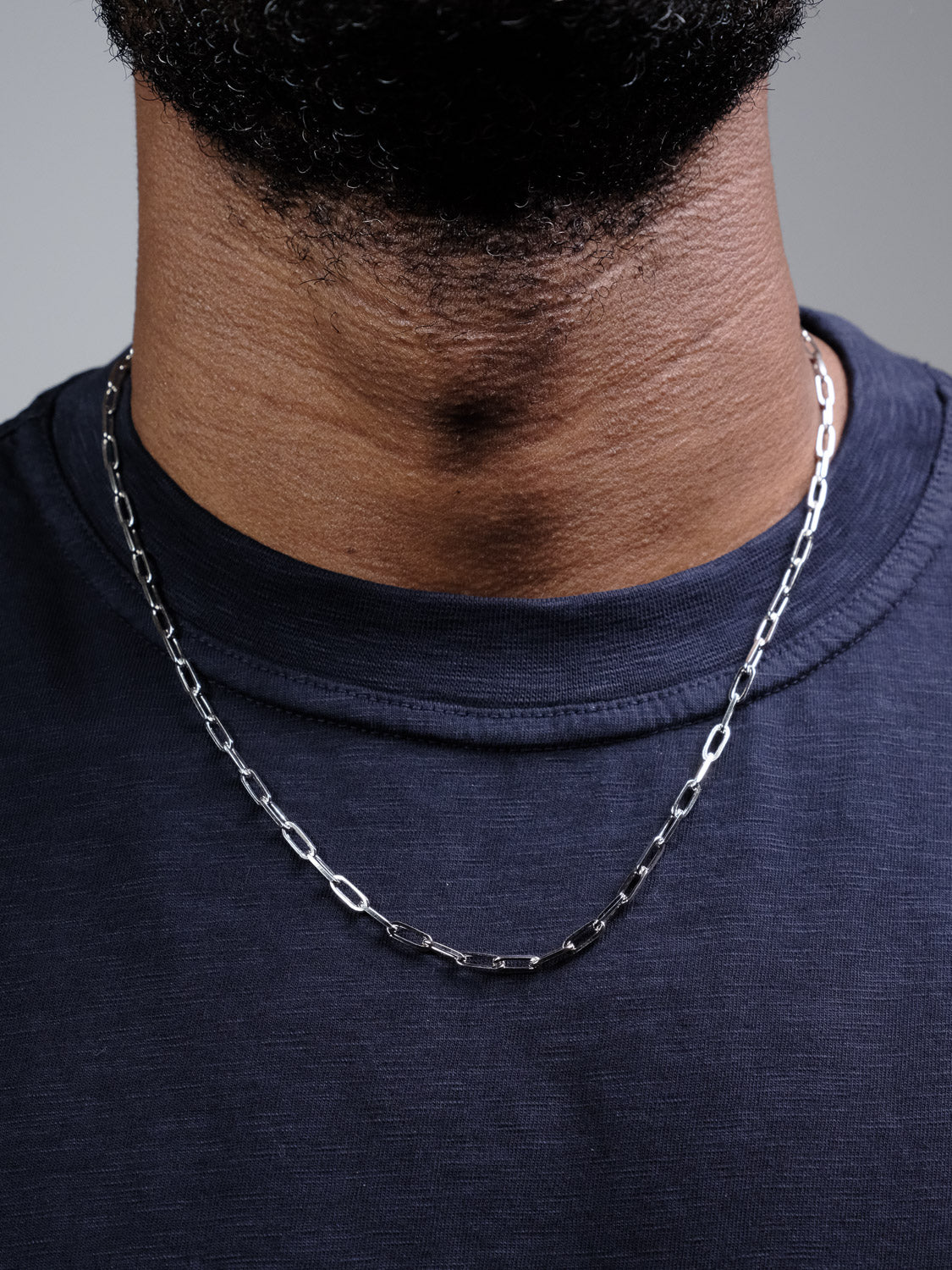 A close-up of a medium-brown-skinned person wearing the Kai 100 Paper Clip Chain Necklace by ID Menswear over a dark navy crew-neck shirt, set against a neutral background.