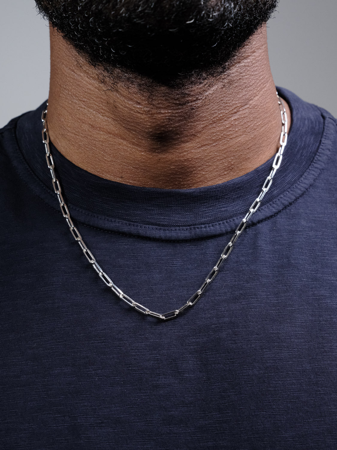 A close-up of a person wearing a navy blue shirt and the Kai 120 Paper Clip Chain Necklace by ID Menswear, showing only the lower half of their face, neck, and upper chest.