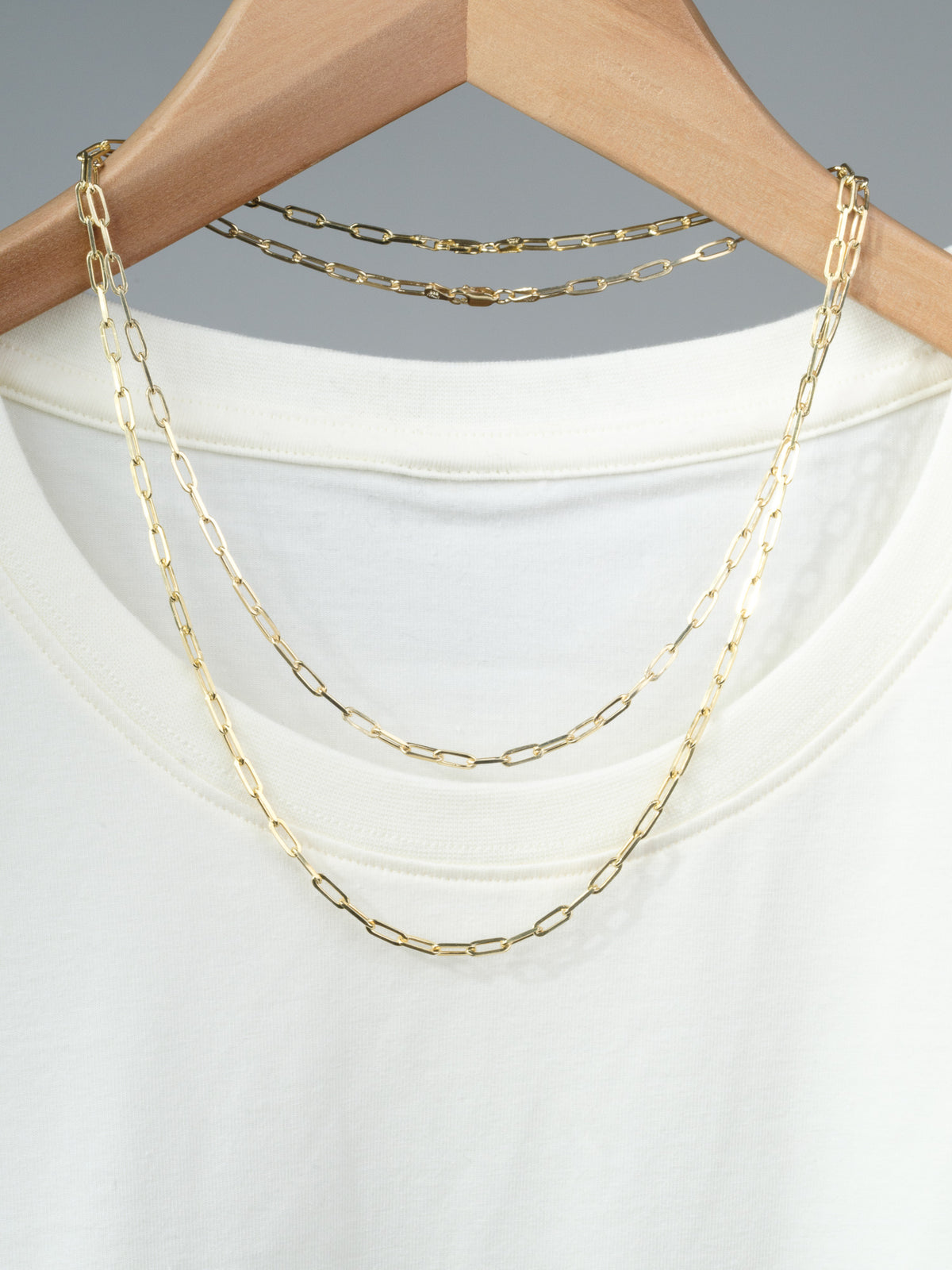 The Kai 100 Paper Clip Chain Necklace by ID Menswear, featuring elongated gold-plated links, hangs on a wooden hanger and is displayed over a plain white T-shirt.