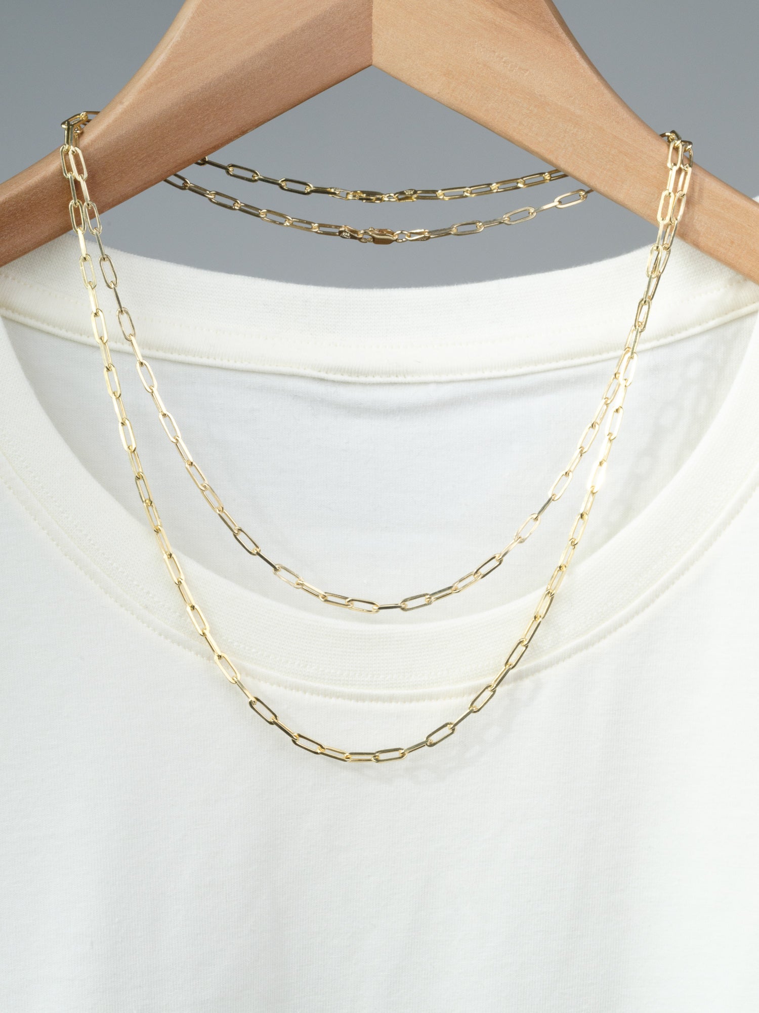 The Kai 100 Paper Clip Chain Necklace by ID Menswear, featuring elongated gold-plated links, hangs on a wooden hanger and is displayed over a plain white T-shirt.