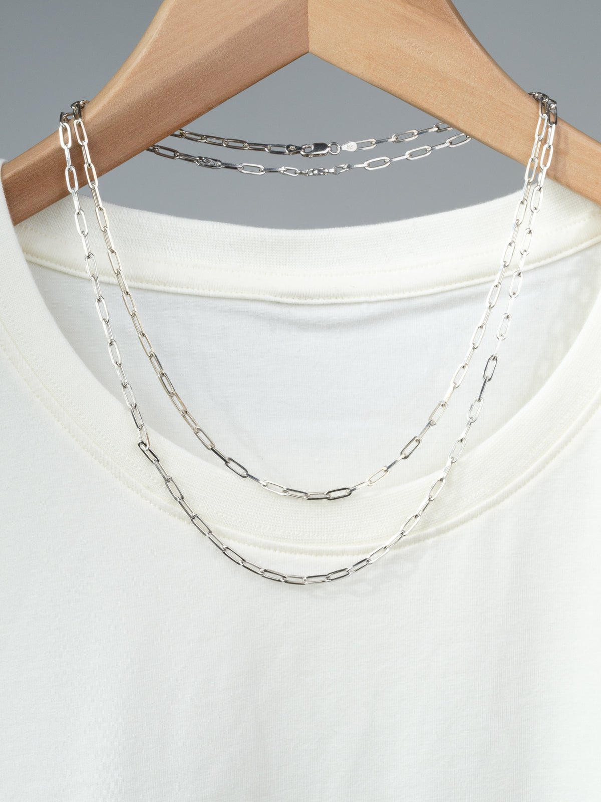 A white t-shirt on a wooden hanger displays two layered Kai 100 Paper Clip Chain necklaces by ID Menswear draped over the collar, set against a neutral background.