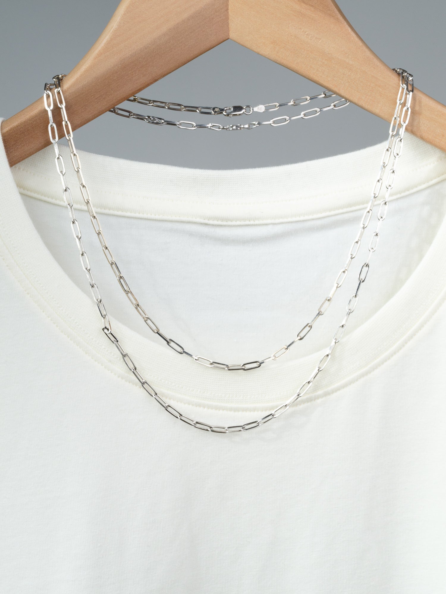 A white t-shirt on a wooden hanger displays two layered Kai 100 Paper Clip Chain necklaces by ID Menswear draped over the collar, set against a neutral background.
