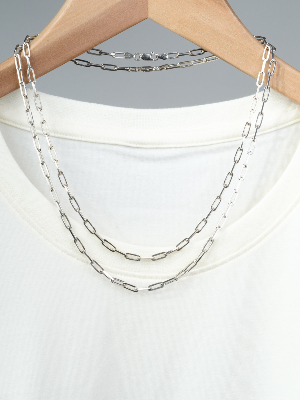 A white T-shirt on a wooden hanger displays two ID Menswear Kai 120 Paper Clip Chain Necklaces, each with elongated rectangular sterling silver links. The background is plain gray.