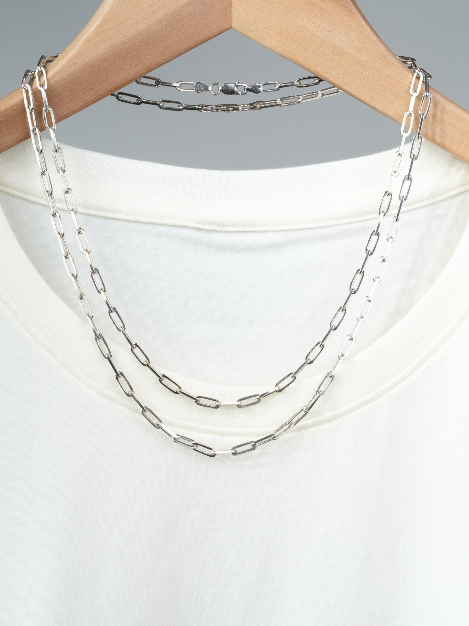 A white T-shirt on a wooden hanger displays two ID Menswear Kai 120 Paper Clip Chain Necklaces, each with elongated rectangular sterling silver links. The background is plain gray.