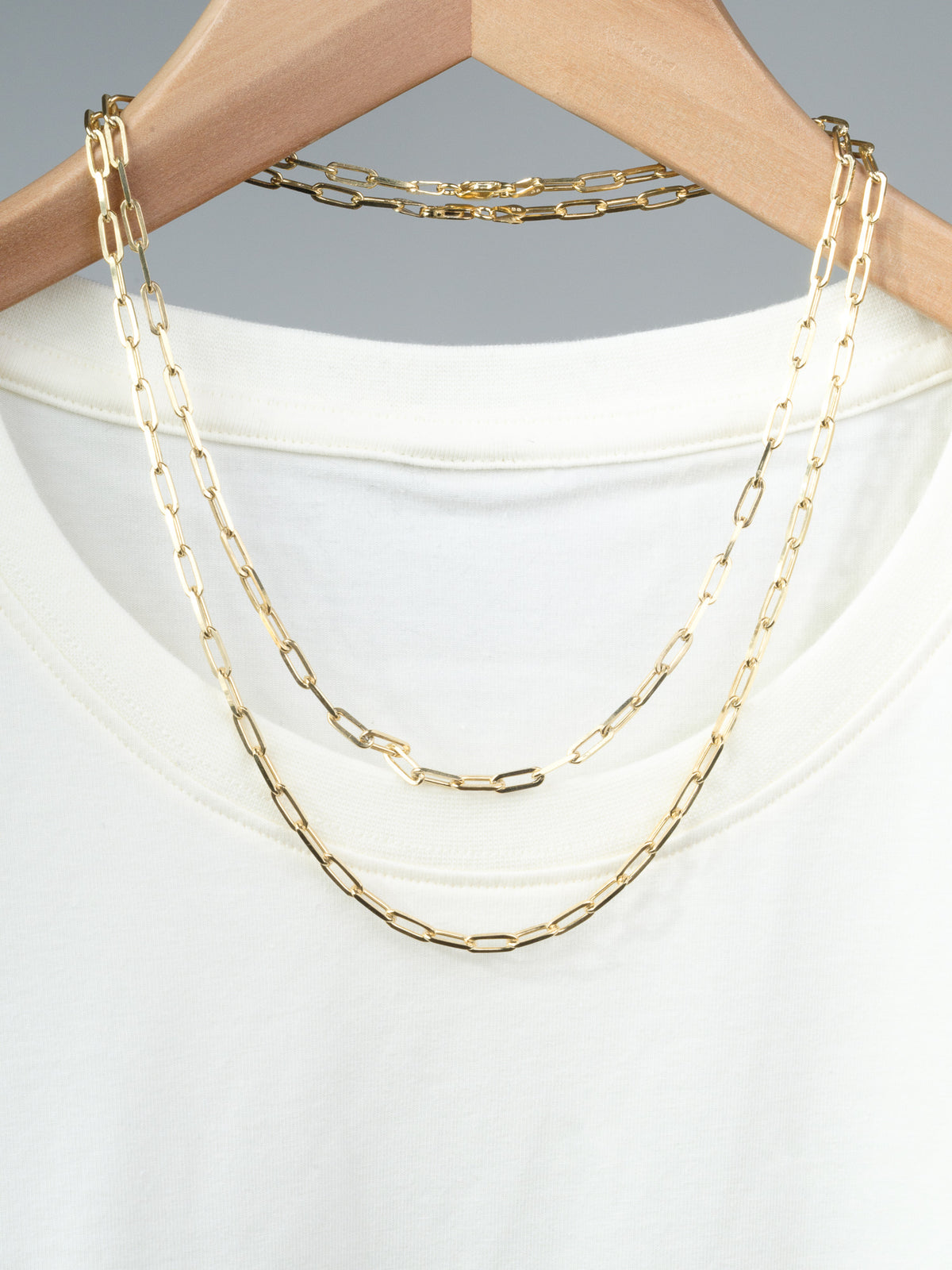 A white T-shirt on a wooden hanger with two layered ID Menswear Kai 120 Paper Clip Chain Necklaces draped over it, set against a plain gray background.