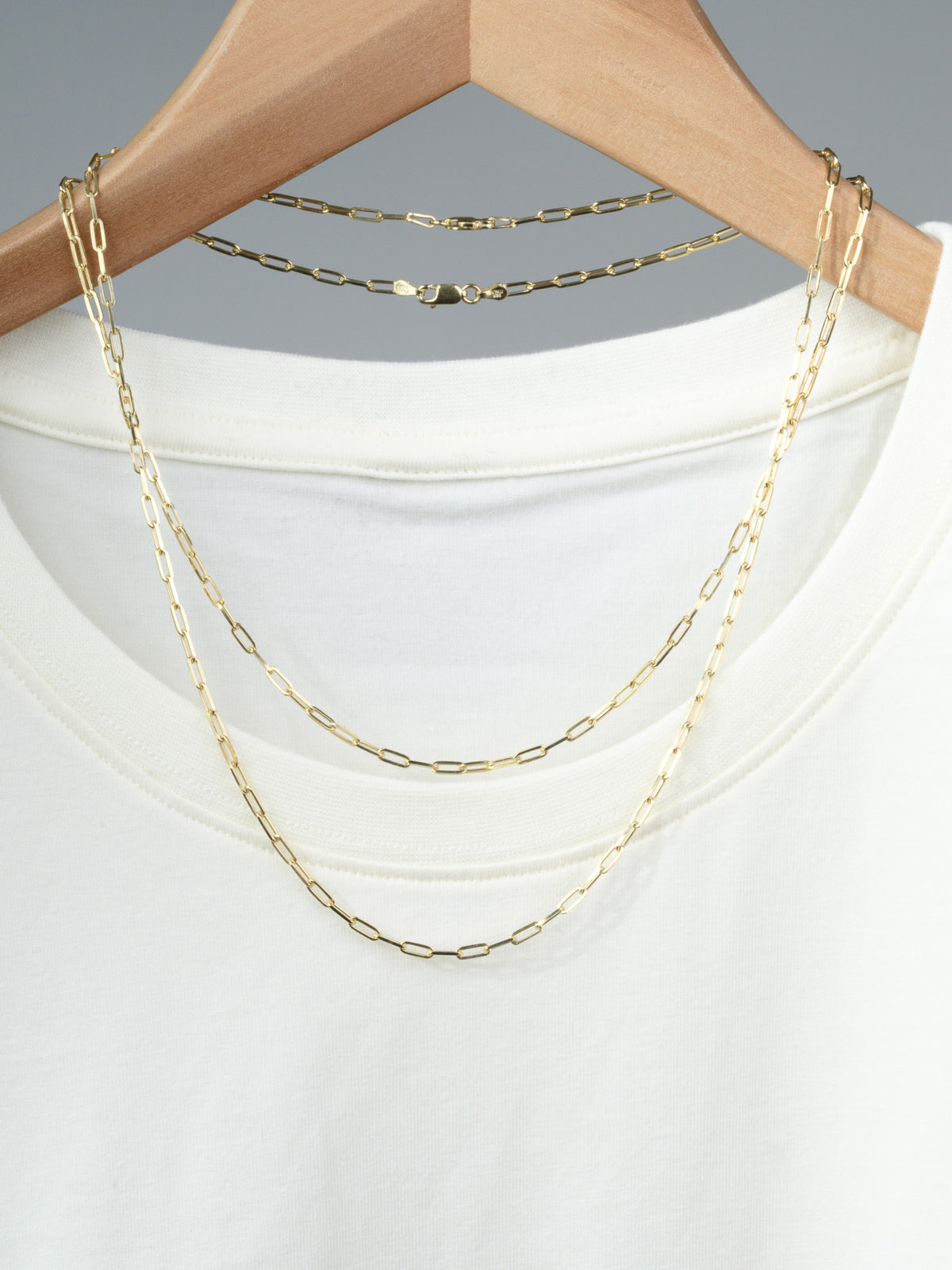 The Kai 80 Paper Clip Necklace by ID Menswear, featuring gold-plated elongated links, is draped over a plain white t-shirt on a wooden hanger against a neutral gray background.