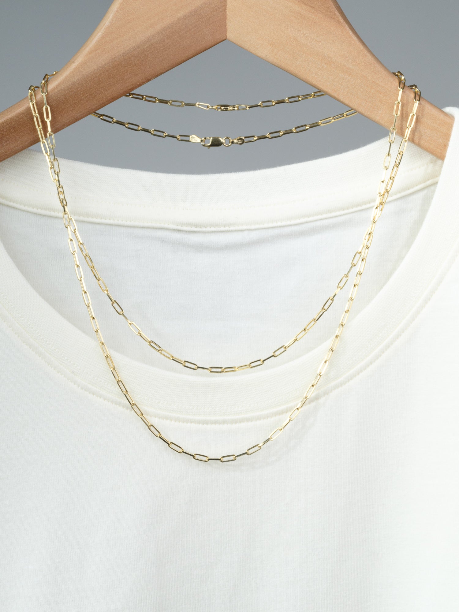 The Kai 80 Paper Clip Necklace by ID Menswear, featuring gold-plated elongated links, is draped over a plain white t-shirt on a wooden hanger against a neutral gray background.