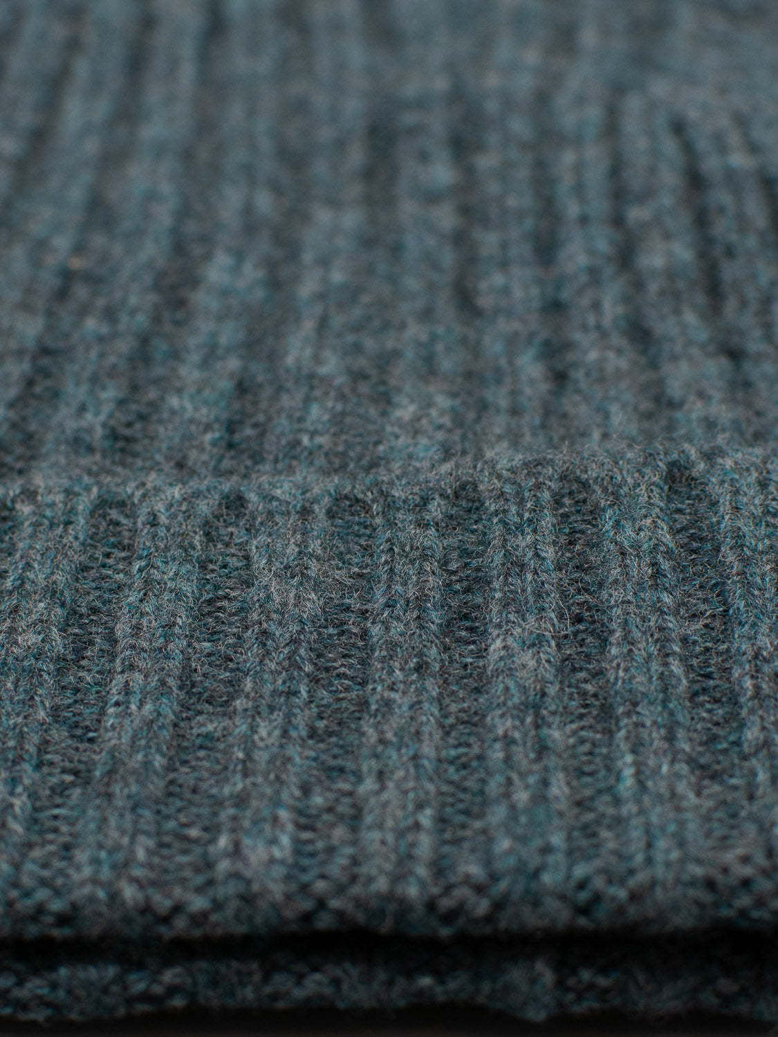 A close-up of Kailash Merino Wool Beanie by ID Menswear showcases the textured, ribbed teal fabric. The material exudes warmth and softness, perfect for cozy outdoor adventures. Intricate patterns and thread details highlight its quality craftsmanship.