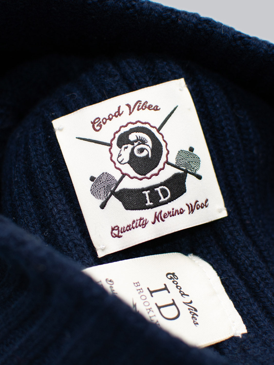Close-up of a clothing label on dark fabric, showcasing a sheep logo with crossed knitting needles. The text reads ID Menswear Kailash Merino Wool Beanie—perfect for outdoor adventures. Another smaller tag says ID Menswear. Enjoy warmth and softness for your Merino Wool Beanie moments.