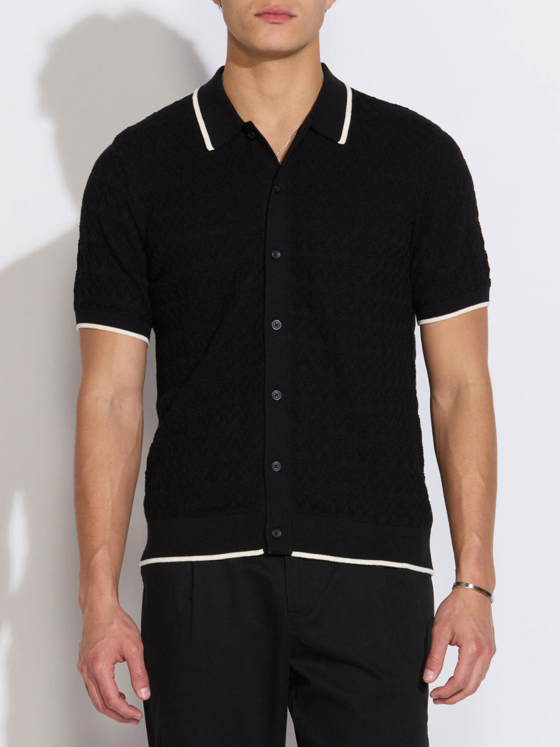 A person in the Kalino Full Placket Sweater Polo by Civil Society—black with white trim on collar, cuffs, and hem—paired with black pants stands against a plain light backdrop, showcasing modern men’s style.