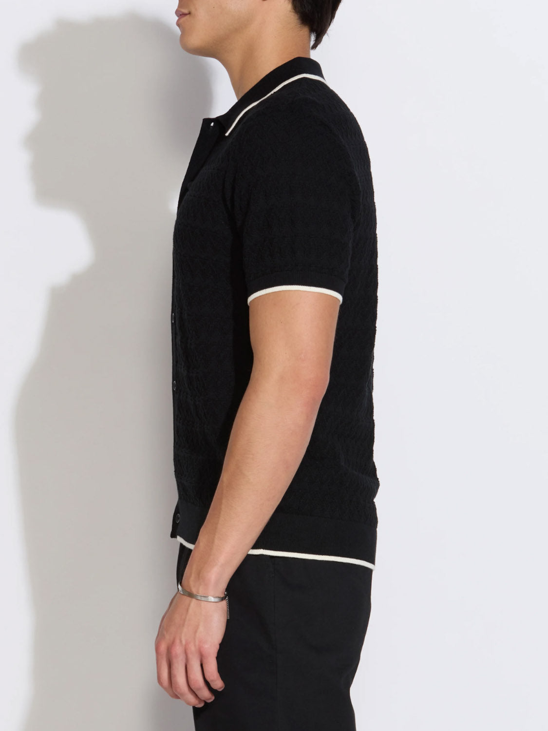 A person in profile stands against a white background, wearing the Civil Society Kalino Full Placket Sweater Polo—black with white trim at the collar, sleeves, and hem—paired with black pants. They have short dark hair and a bracelet on their left wrist.