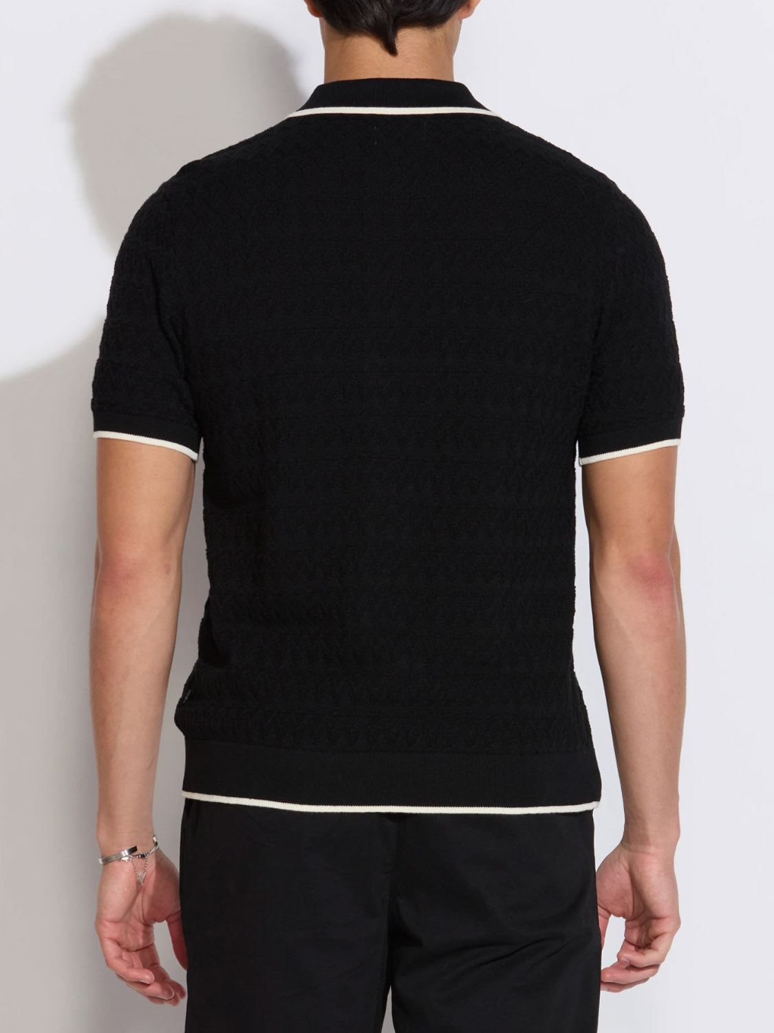 A person faces away from the camera, wearing Civil Society's Kalino Full Placket Sweater Polo—a short-sleeve black textured polo with white trim on the sleeves, collar, and hem—paired with black pants for an elevated men's look.