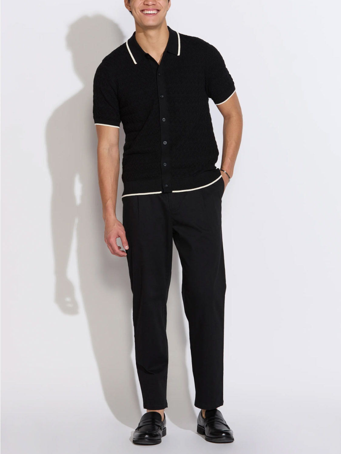 A person stands against a white background in the Civil Society Kalino Full Placket Sweater Polo, paired with black trousers and dress shoes—an effortlessly sharp, modern look. Smiling, one hand in their pocket.