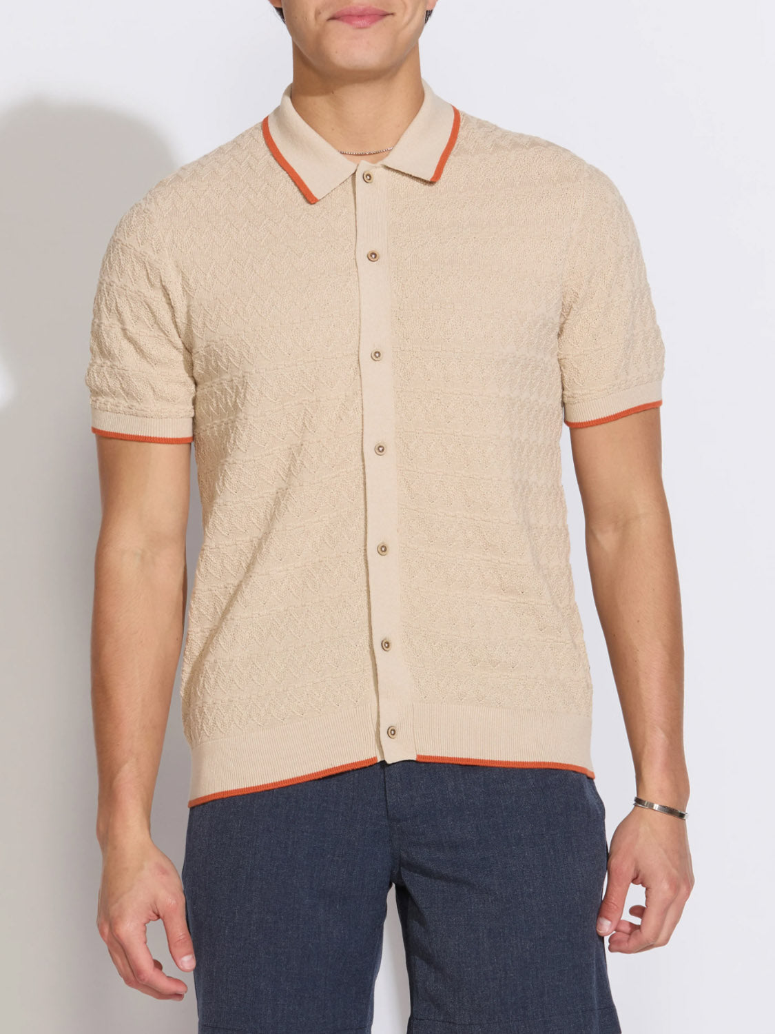 A man models the Civil Society Kalino Full Placket Sweater Polo in beige with orange trim and dark blue pants against a plain white background, showcasing effortless modern men's style.