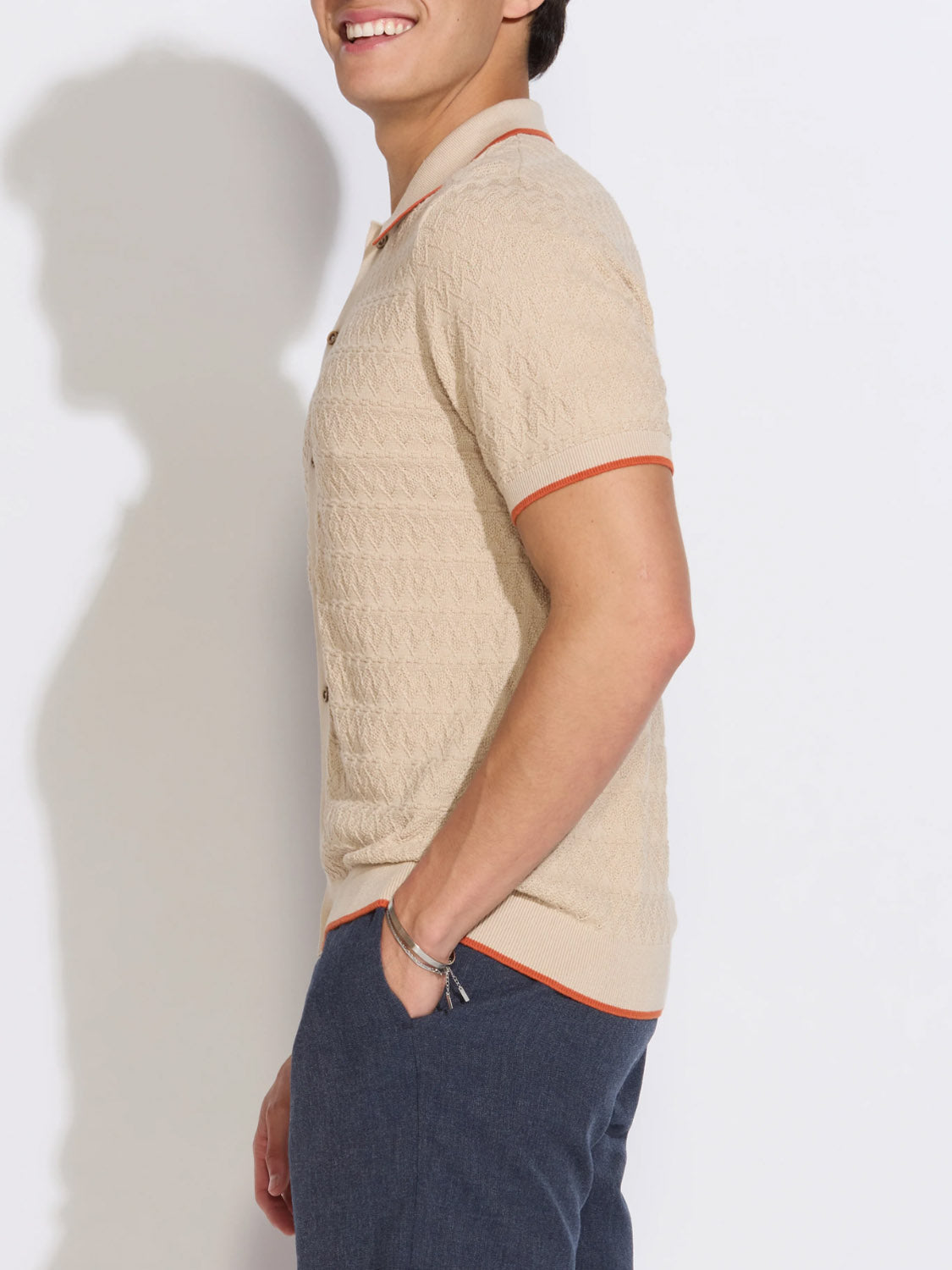 A man smiles in profile, wearing the Civil Society Kalino Full Placket Sweater Polo in beige with orange trim, paired with blue pants and a silver bracelet—a stylish take on men's fashion.