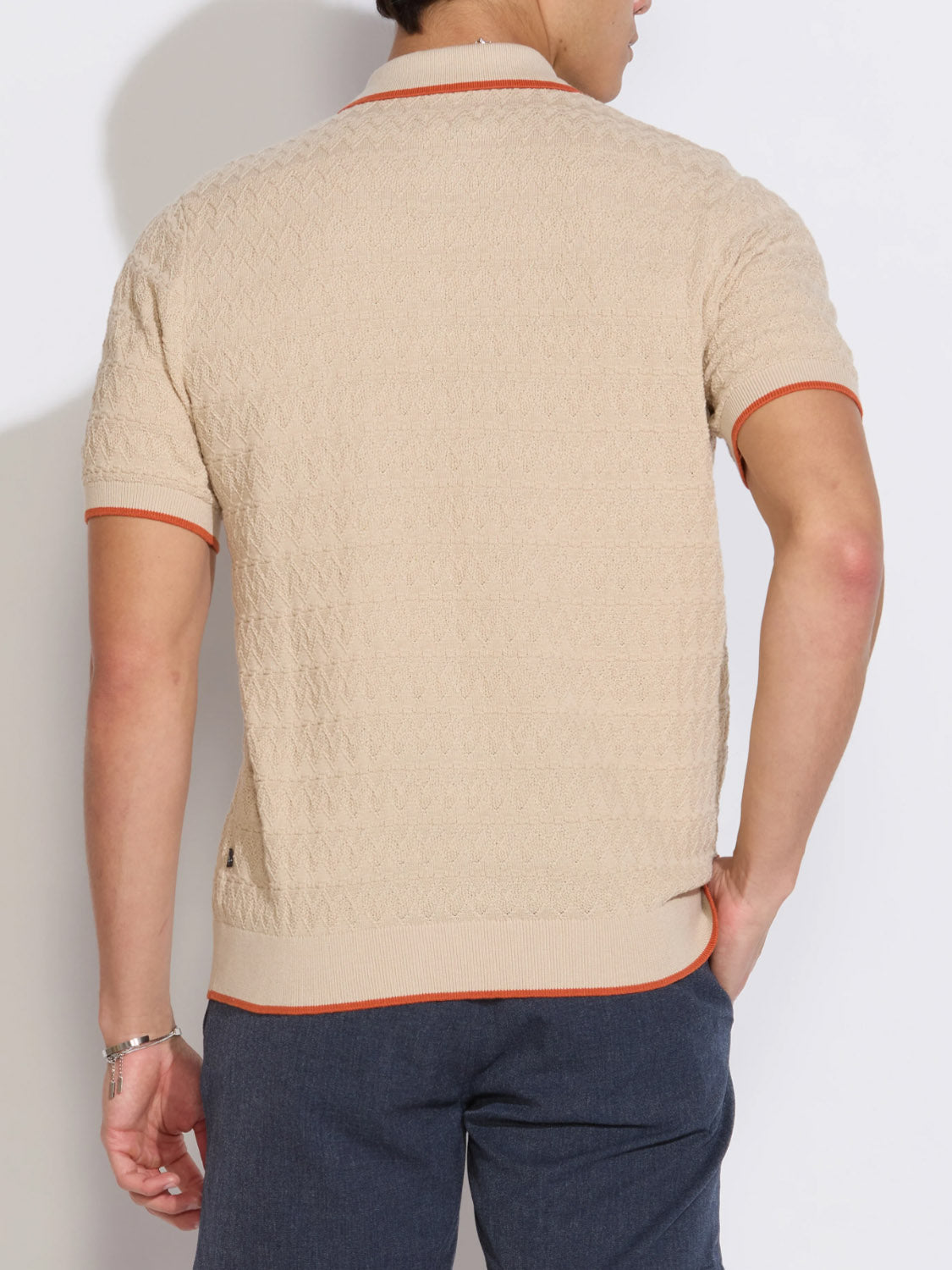 A man stands with his back to the camera, wearing the Civil Society Kalino Full Placket Sweater Polo—a textured beige style with orange trim—paired effortlessly with blue pants for a modern, stylish look.