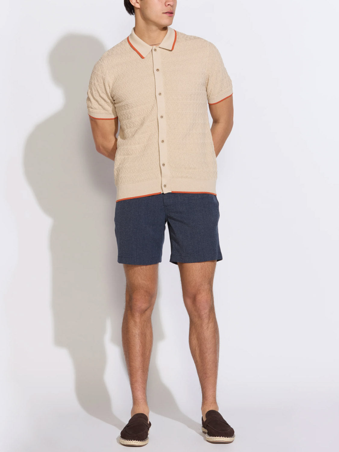 A man showcases effortless men's fashion in the Civil Society Kalino Full Placket Sweater Polo, paired with dark blue shorts and brown slip-on shoes, posing with his hands behind his back against a plain light background.