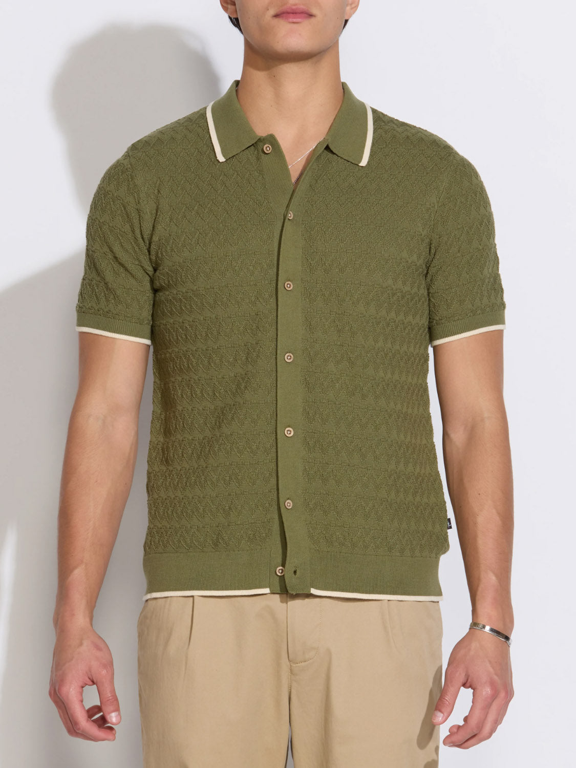 A man wears the Kalino Full Placket Sweater Polo by Civil Society—a short-sleeve, olive green collared top with beige trim—paired with beige pants, standing against a plain light background for an effortless style statement.
