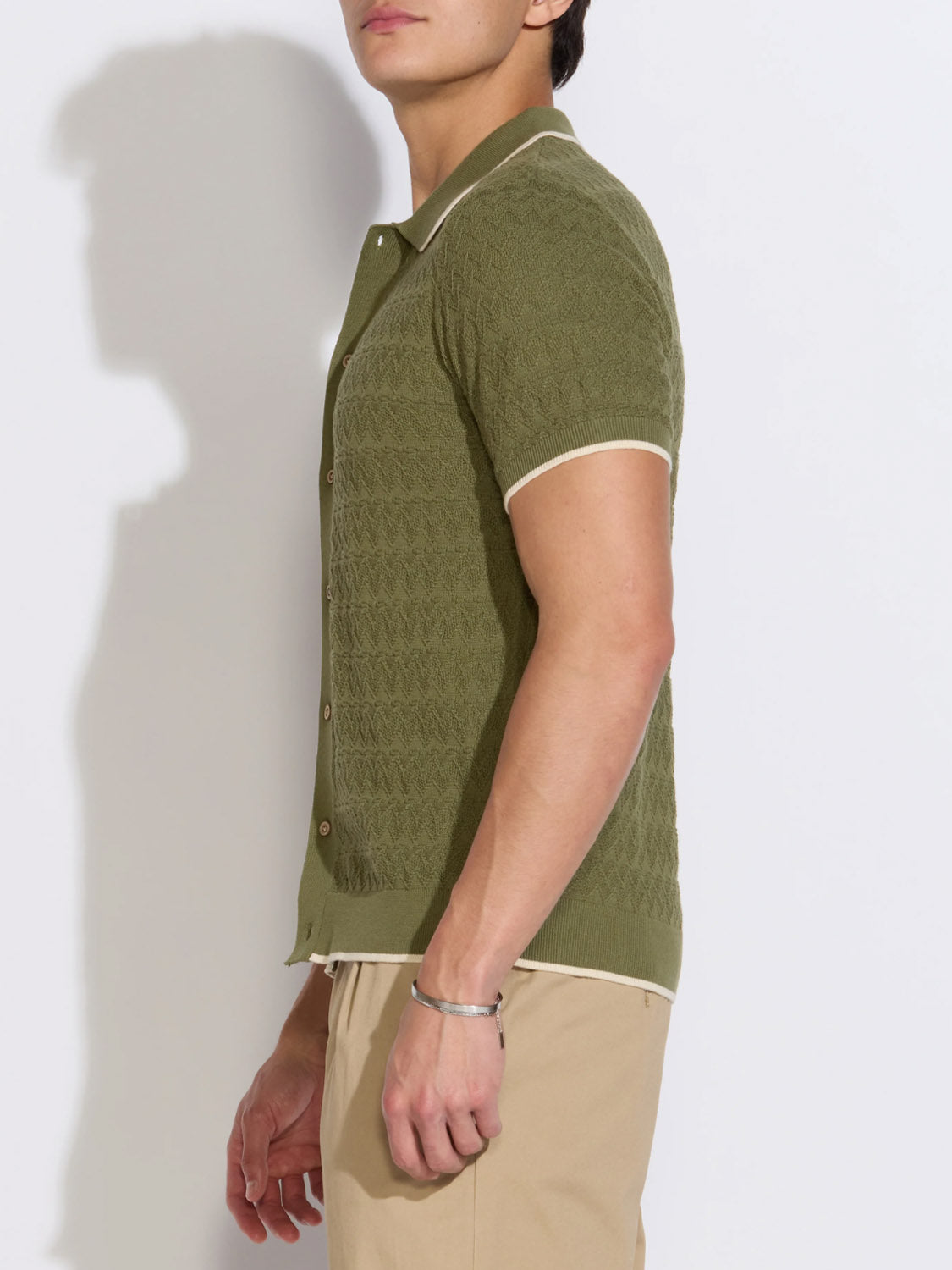 Wearing Civil Society's Kalino Full Placket Sweater Polo in olive green with beige trim and beige pants, a person stands sideways, head turned slightly away from the camera.