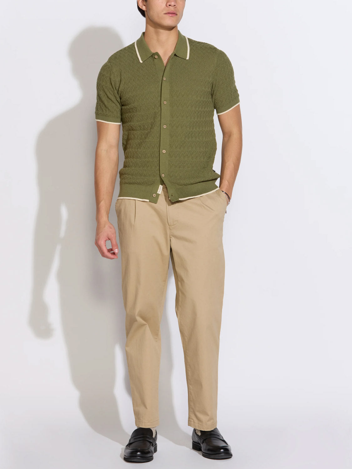 A man models Civil Society's Kalino Full Placket Sweater Polo—an olive green short-sleeve shirt with white trim—paired with beige pants and black dress shoes, posing with one hand in his pocket against a plain background.