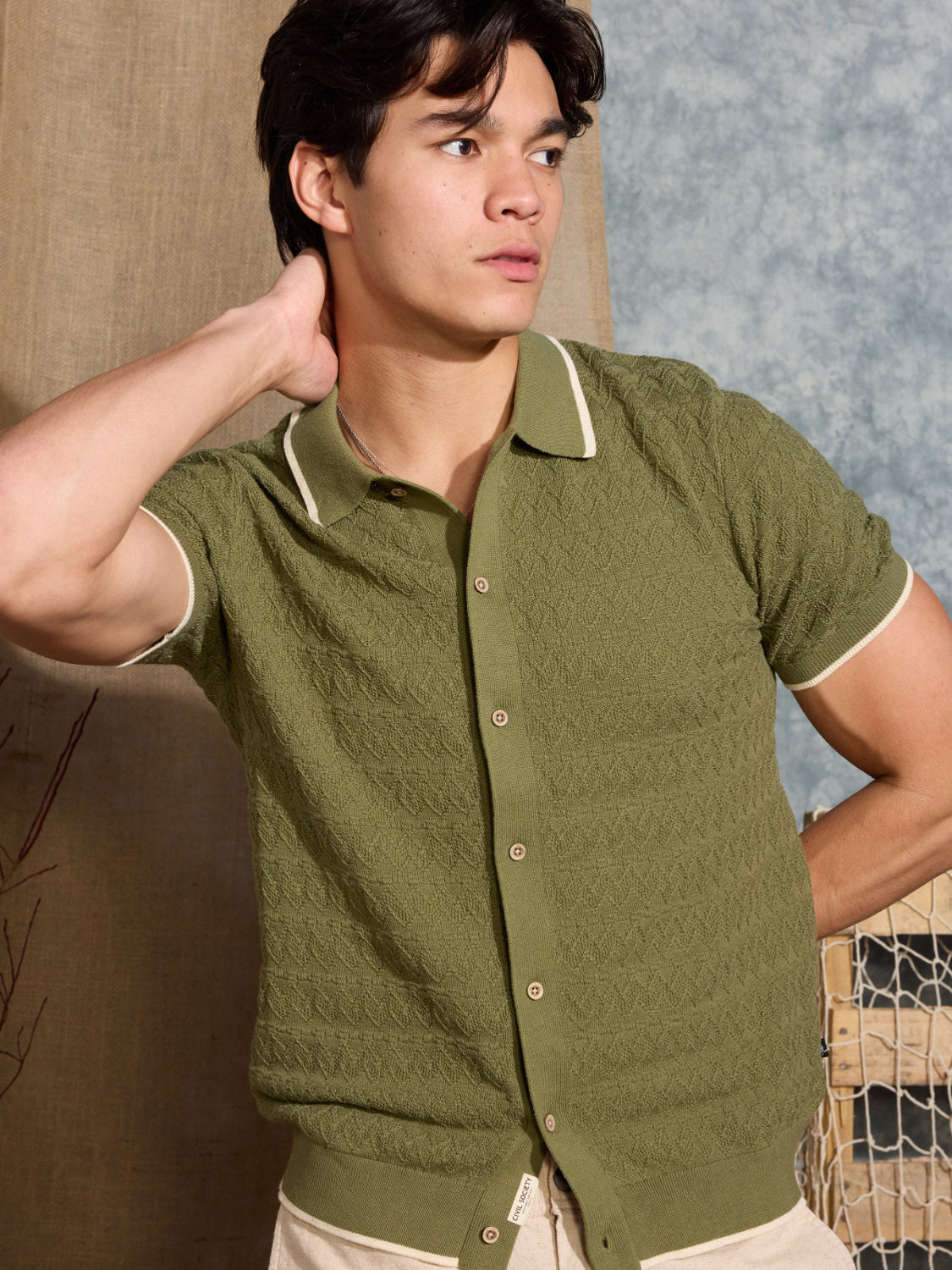 A young man with dark hair models the Civil Society Kalino Full Placket Sweater Polo in textured green with beige trim, standing before a neutral backdrop and gazing to the side with one hand behind his neck.