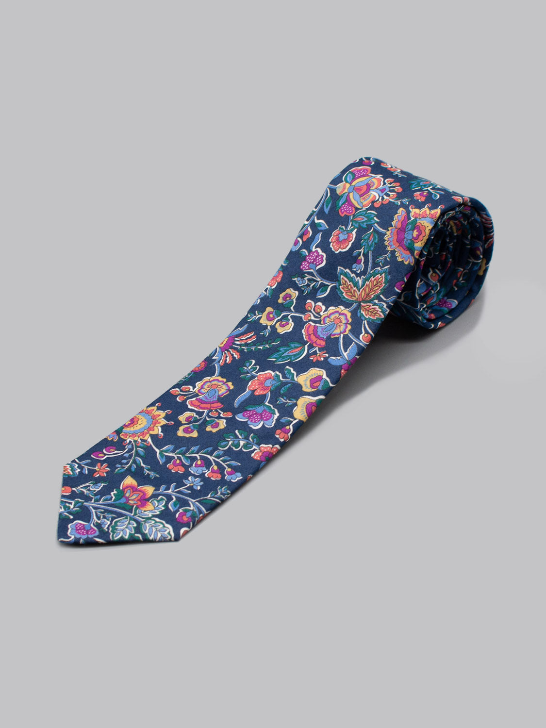 The Liberty of London Tana Lawn™ Kausa Tie by ID Menswear, handmade in Brooklyn from floral-patterned dark blue Tana Lawn cotton, is shown rolled up on a plain gray surface.