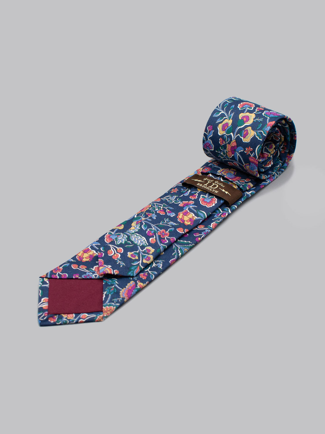 The Liberty of London Tana Lawn™ Kausa Tie by ID Menswear is rolled up and displayed on a plain gray background, featuring a blue base with pink, yellow, and orange floral patterns on premium cotton.