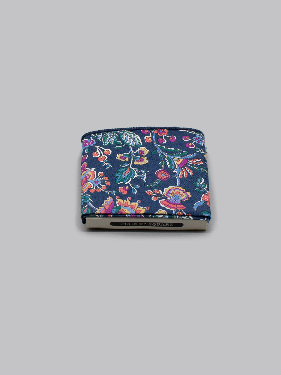 The Liberty™ Kazusa Pocket Square by ID Menswear, made from Liberty of London Tana Lawn cotton with a vibrant floral design on a dark background, is shown folded on a gray surface with the HOUSE OF JULLA label visible along the edge.