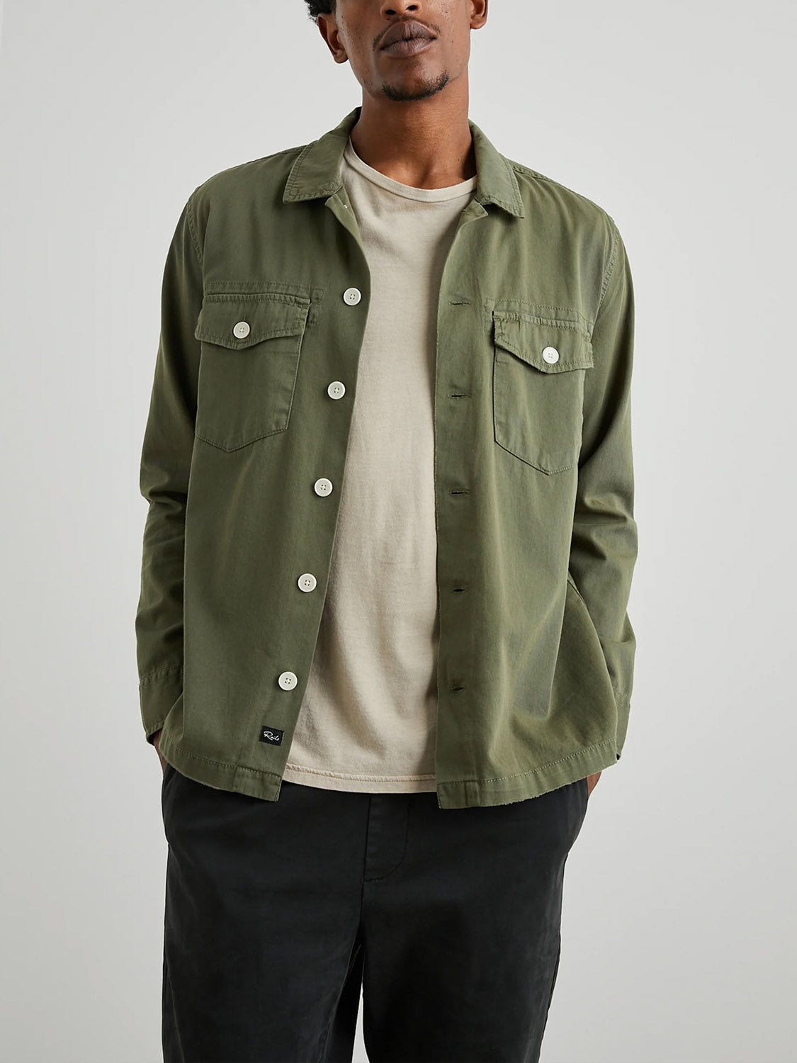 A person wears a Rails Kerouac Shirt Jacket, a classic fit green button-up resembling a military shirt, over a beige t-shirt and black pants. The cotton twill jacket by Rails includes two buttoned chest pockets and stands against a neutral background.