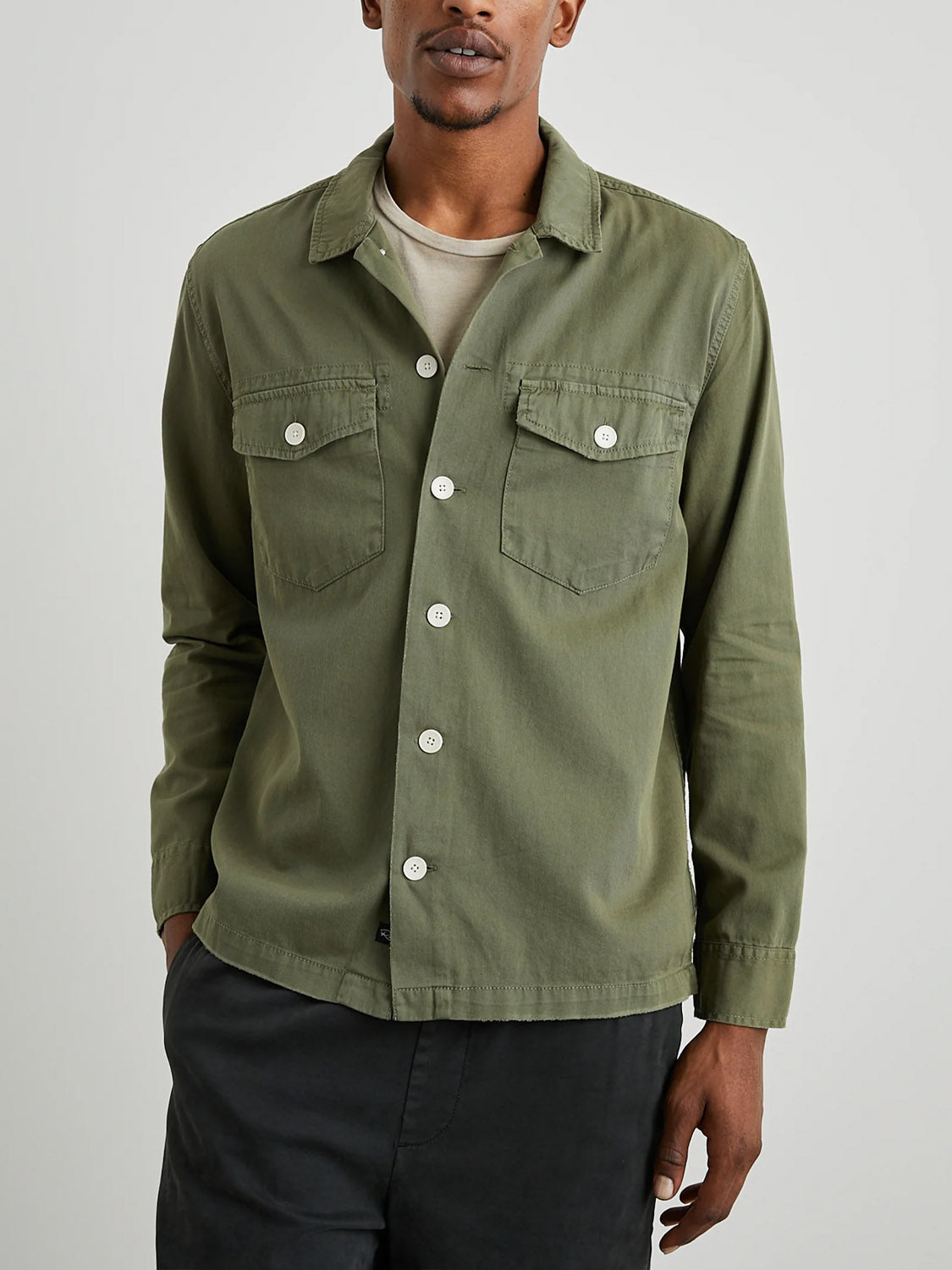 A person is wearing a Rails Kerouac Shirt Jacket in olive green, featuring a classic fit and two chest pockets, over a light beige t-shirt. This ensemble is paired with black pants. They stand against a plain light background with one hand in their pocket.