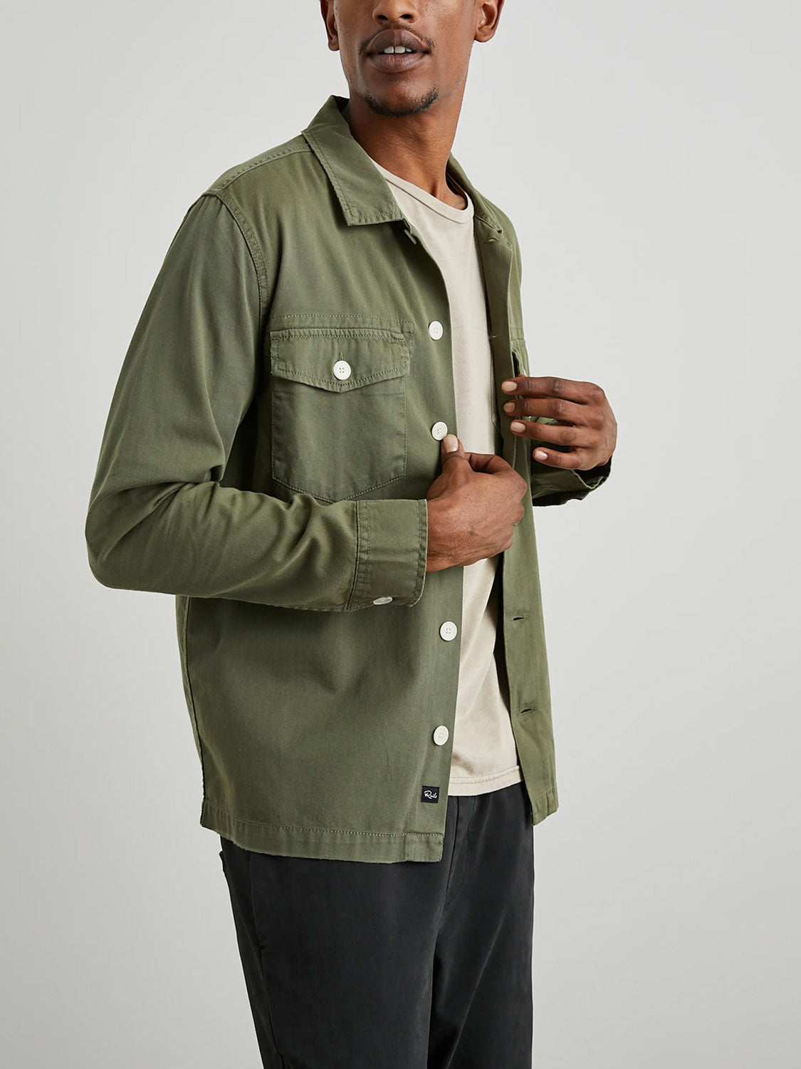 A person models the Rails Kerouac Shirt Jacket in olive green against a plain backdrop. This classic fit features white buttons, a chest pocket, and is buttoned halfway to reveal the beige shirt below as they gaze sideways.