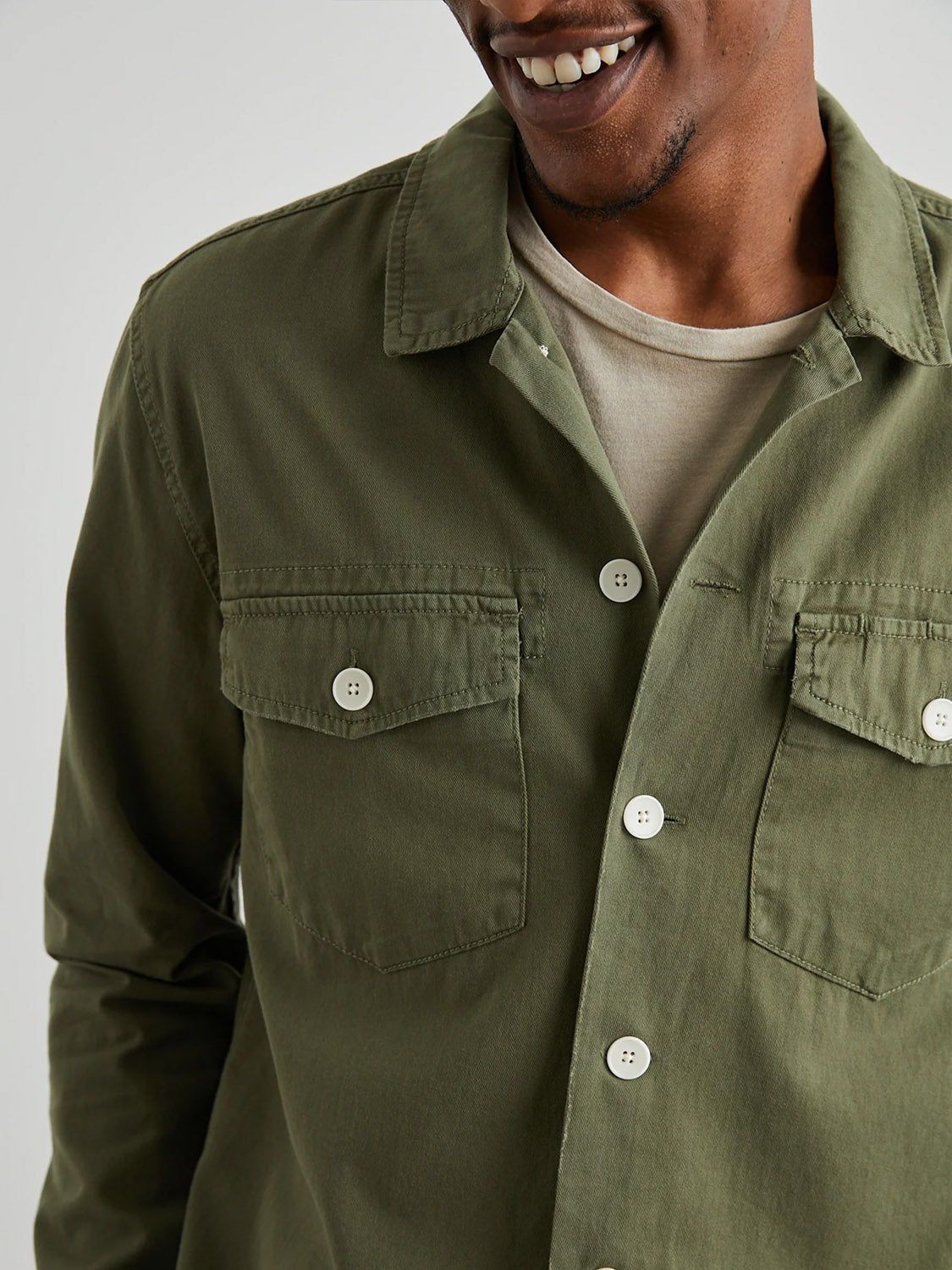 A person is smiling while wearing the Rails Kerouac Shirt Jacket, an olive green, classic-fit military-style shirt jacket over a beige shirt. The cotton twill design includes two flap chest pockets and large white buttons against a light gray background.