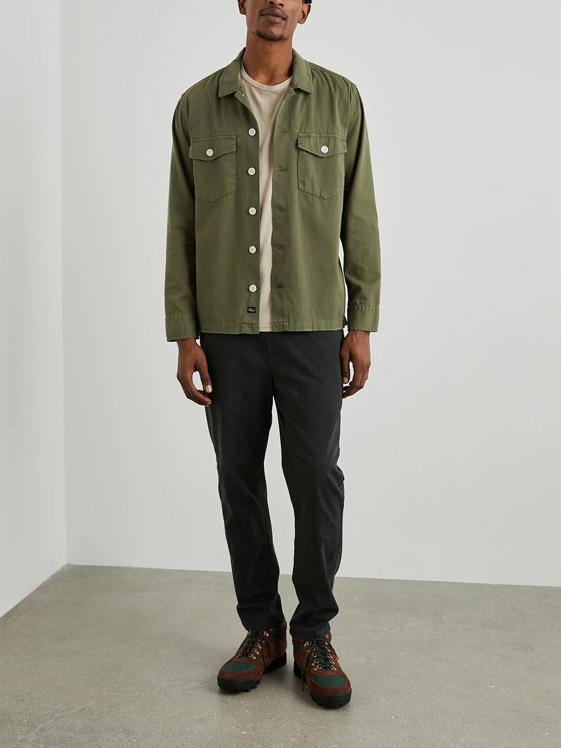 A person leans casually against a white wall in a Rails Kerouac Shirt Jacket over a beige tee, pairing it with black pants and brown hiking boots. Hands in pockets, they exude classic fit confidence on the light gray concrete floor.