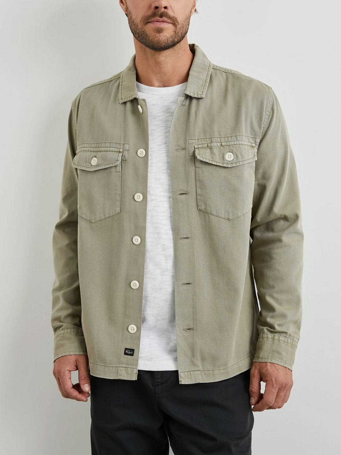 A man wearing the Rails Kerouac Shirt Jacket in light olive-green with two chest pockets over a white t-shirt and black pants, standing against a plain light-colored wall.